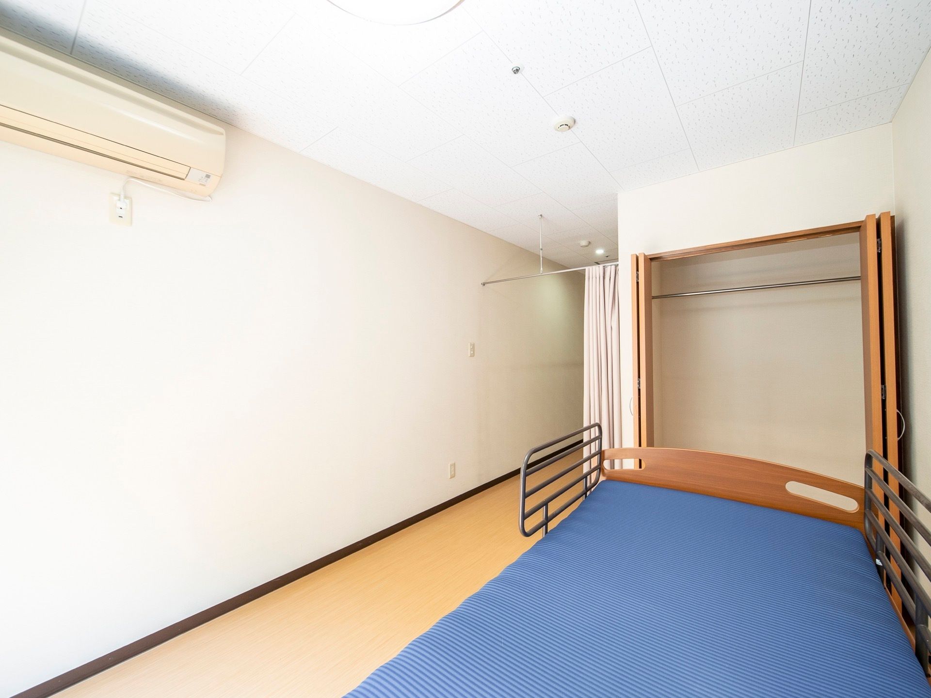 白い壁とライトブラウンの床の居室である。介護用ベッドが設置されている。青いマットレスが敷かれ、柵が付いている。