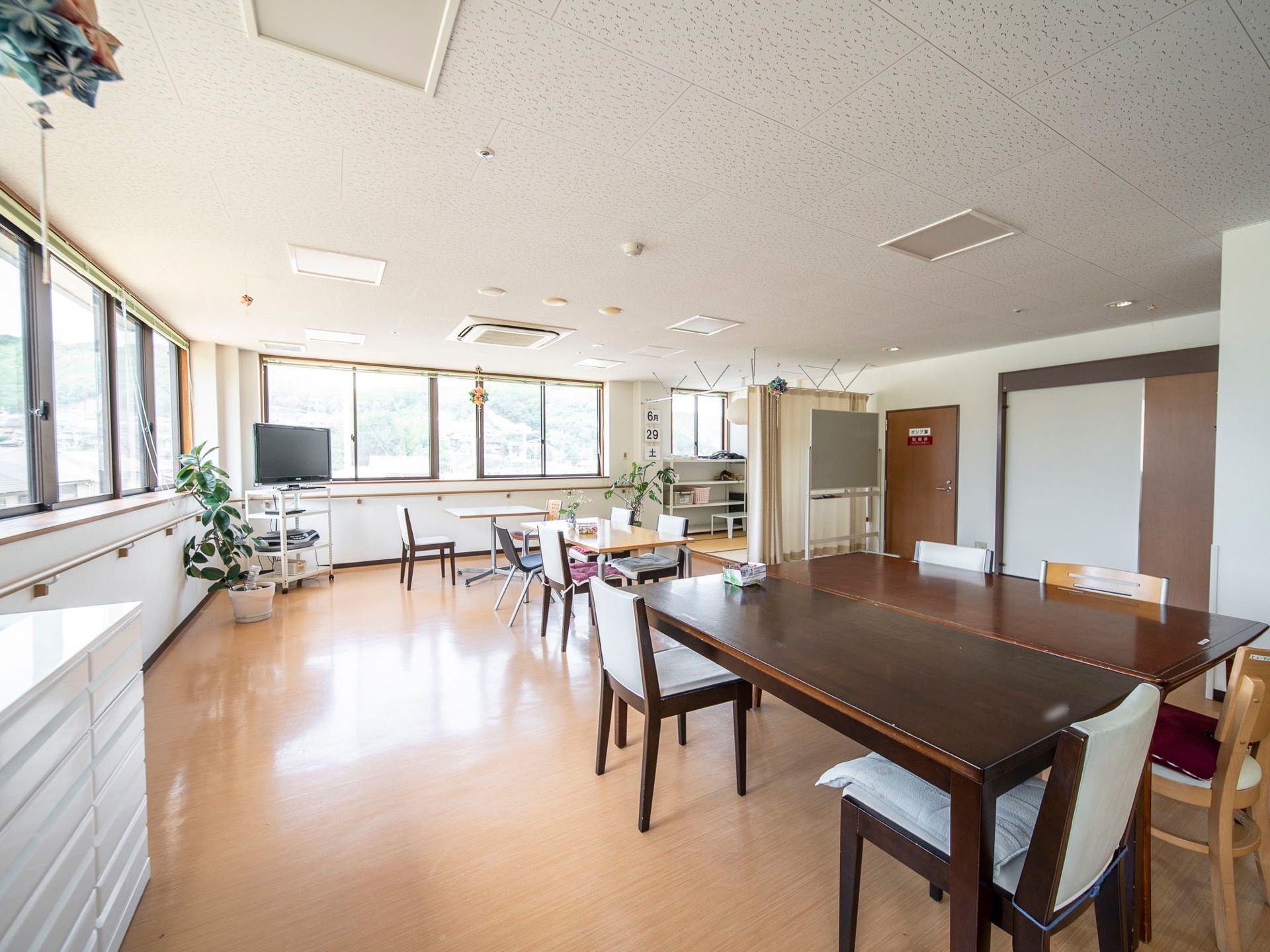 大きなテーブルと椅子が並べられている食堂である。大きな窓が並んでおり、壁には手すりが設置されている。テレビと観葉植物が置かれている。