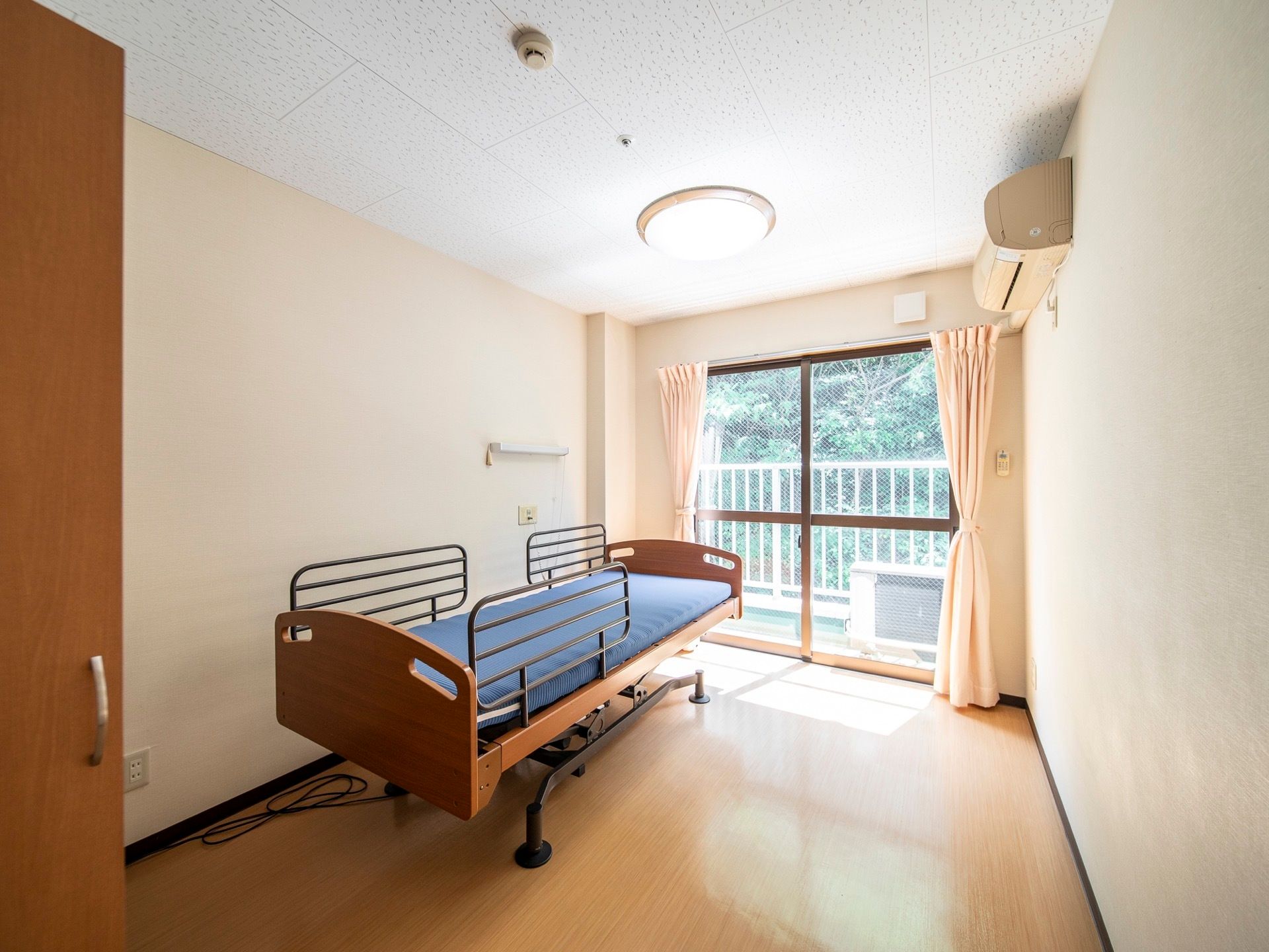 居室には大きな窓が設置され、明るい。青いマットレスの置かれた介護用ベッドがある。窓の外はベランダがあり、緑が生い茂っている。
