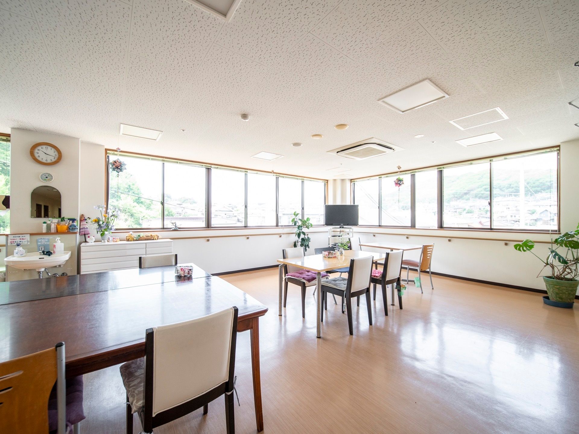 壁一面に窓が並んでいる角部屋の食堂である。テーブル、椅子、テレビ、棚が置かれている。鏡と洗面台もある。
