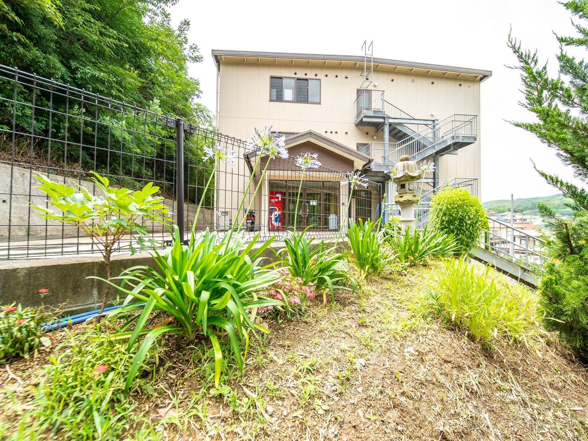 高台にある３階建ての建物には、外付けの非常階段がある。周囲は崖になっており、柵が付いている。建物周辺には木々が立っている。