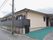 クリーム色の壁面の平屋の建物。道路に面する部分は黒いフェンスが続いている。入り口までは傾斜になっておりグリーンの敷物がされている。
