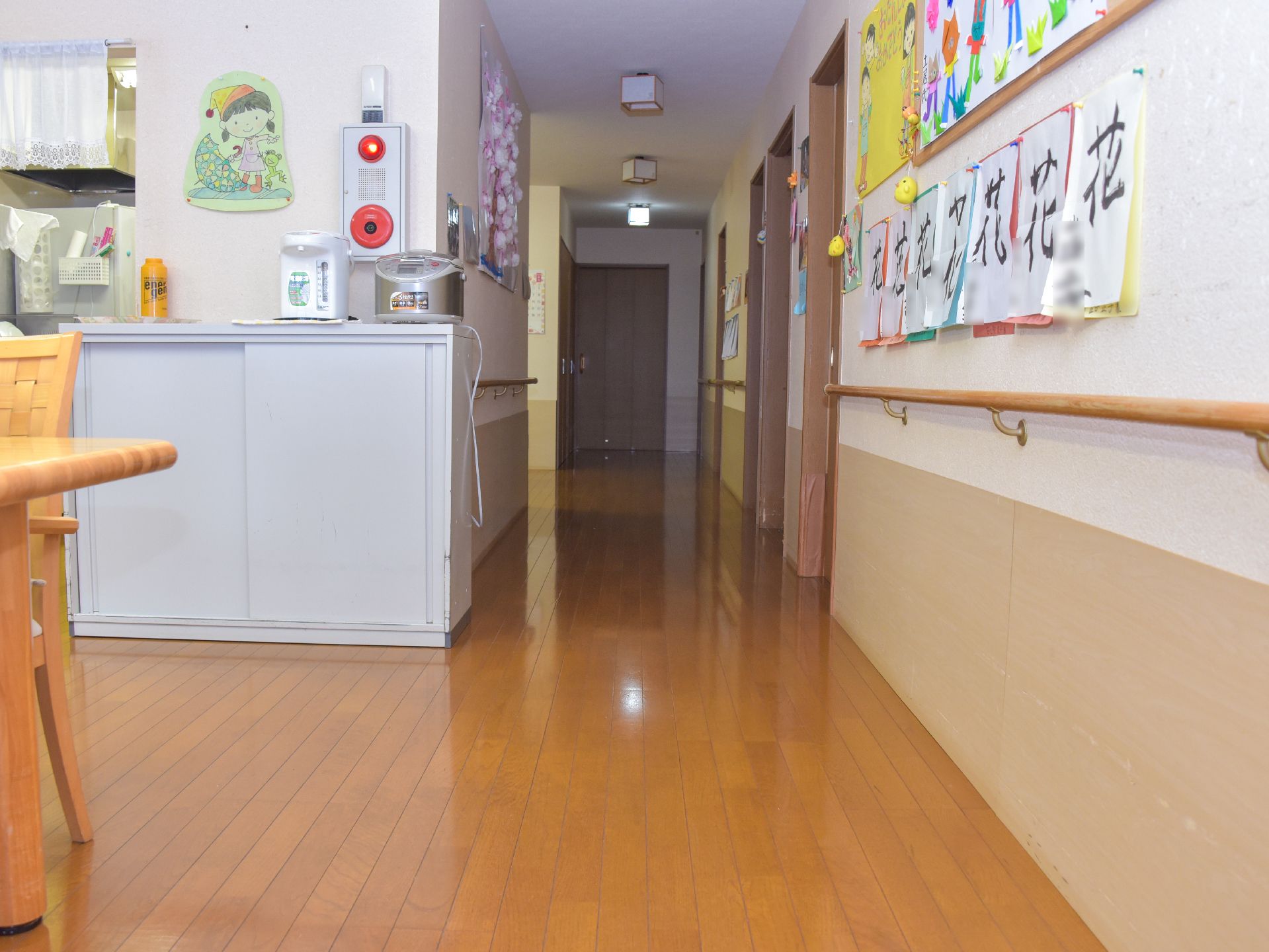 廊下には書や絵などの作品が掲示されている。戸棚の上には炊飯器やポットなども置かれ、壁に非常ベルがある。