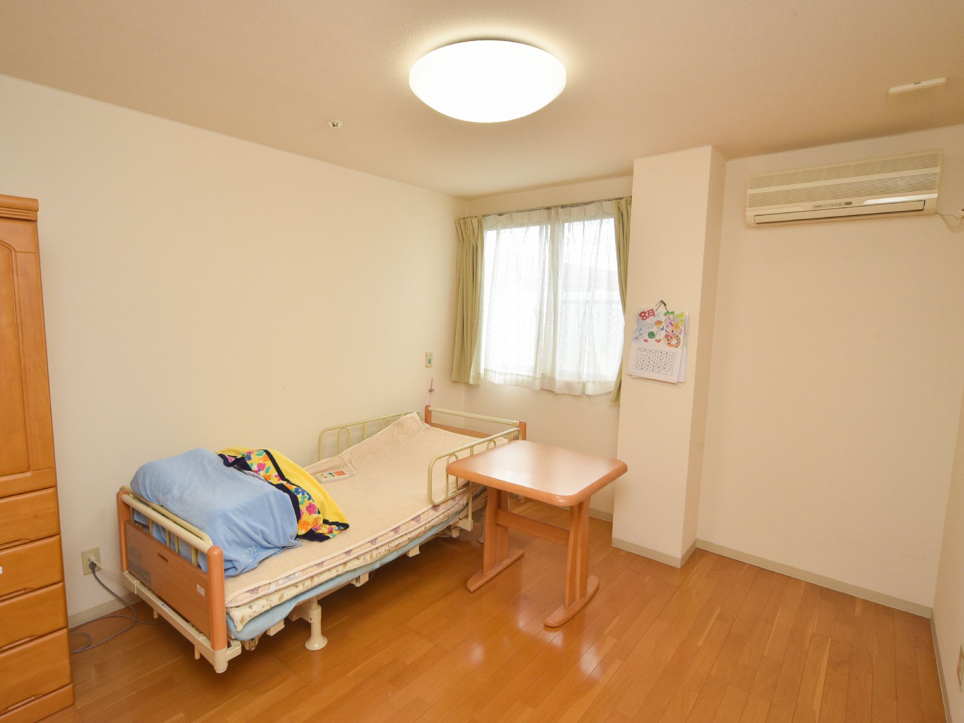 電動式のベッドが置かれていて、枕元には窓があり、外の光がさしこむ。ベッドの脇には小型のテーブルが置かれている。