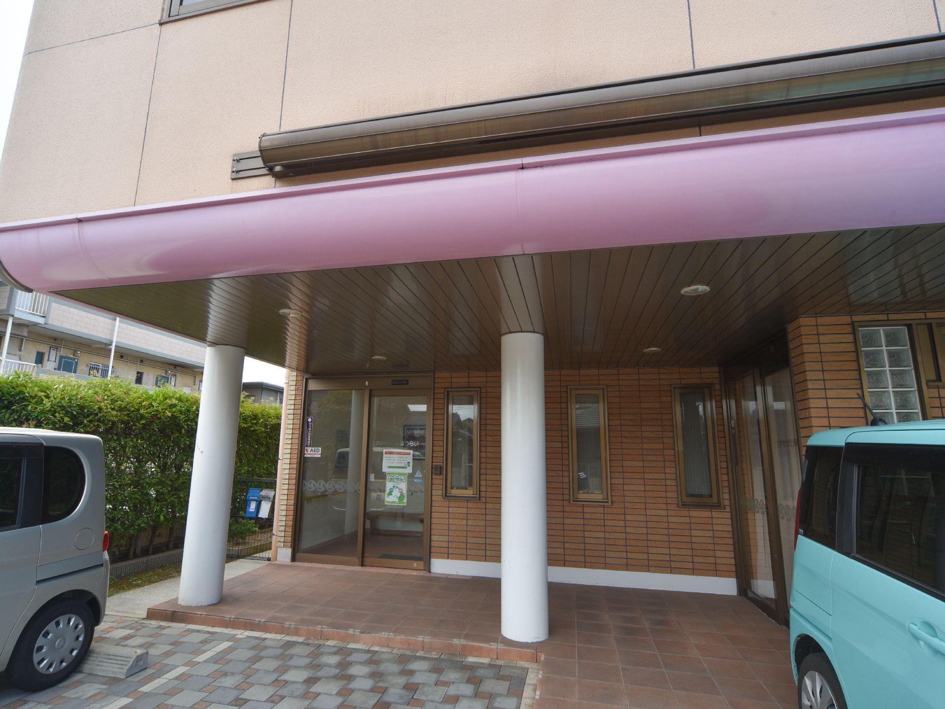 玄関の自動ドア前には2本の白い柱があり、屋根があるスペースが設けられている。その前には駐車場がある。