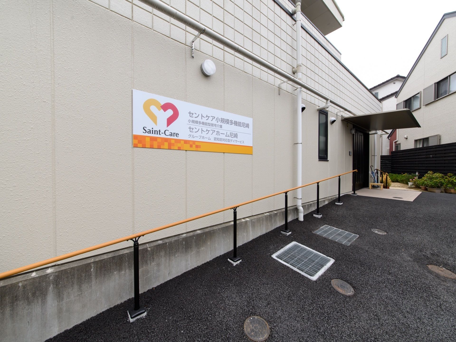 施設の入口には大きな屋根が設置されている。入口の横側の壁には長い手すりが取り付けられている。手すりの上には施設の看板がある。