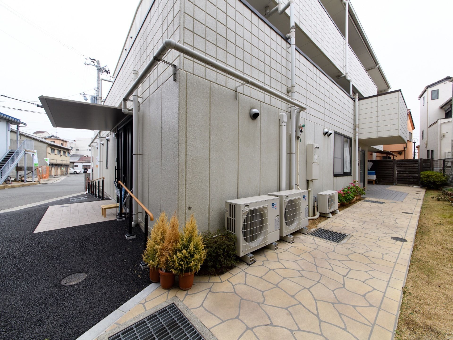 施設の外は舗装されている。室外機が並べられた横には植木鉢が2つ置かれている。施設の入口扉には大きな屋根が設置されている。