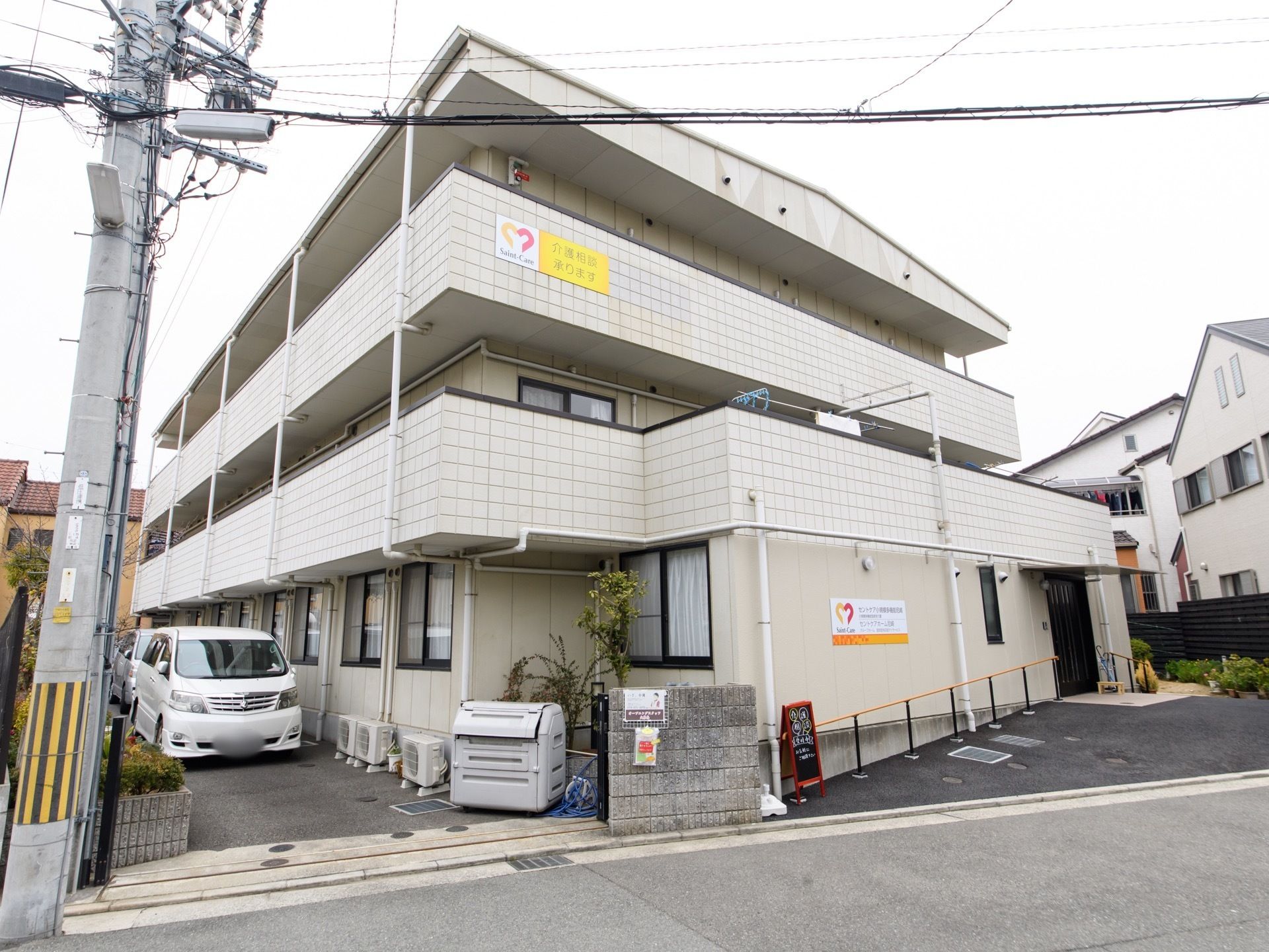 施設は3階建ての造りである。入口の扉横は傾斜があり、手すりが付けられている。施設建物の横側には車が1台止められている。
