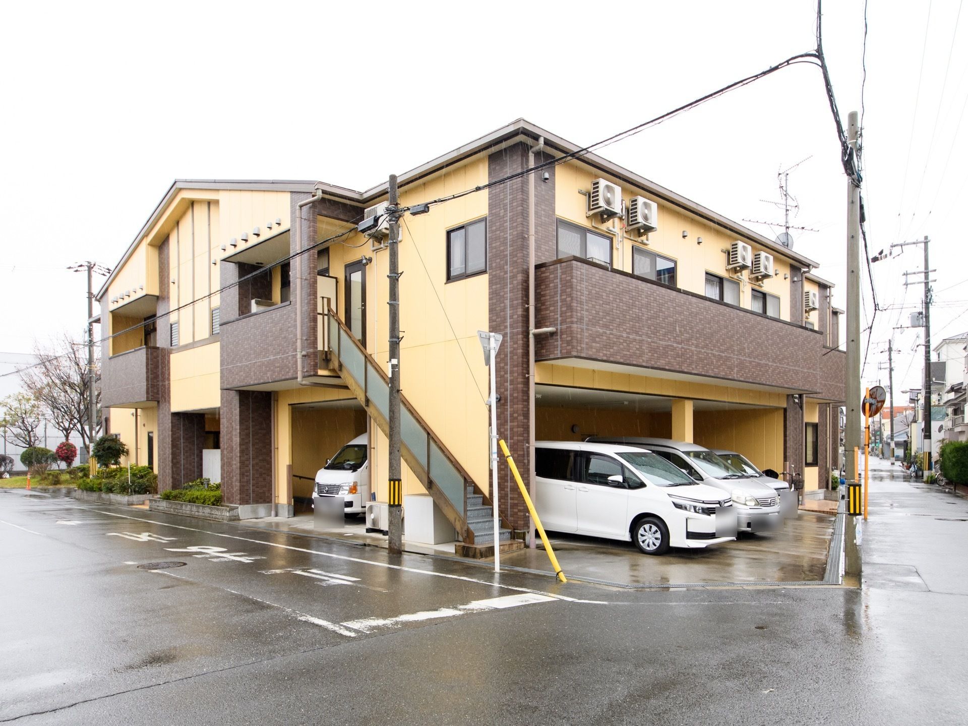 施設は２階建てで黄色と茶色の２色の外観である。１階部分は車の駐車スペースになっていて、車が４台停められている。