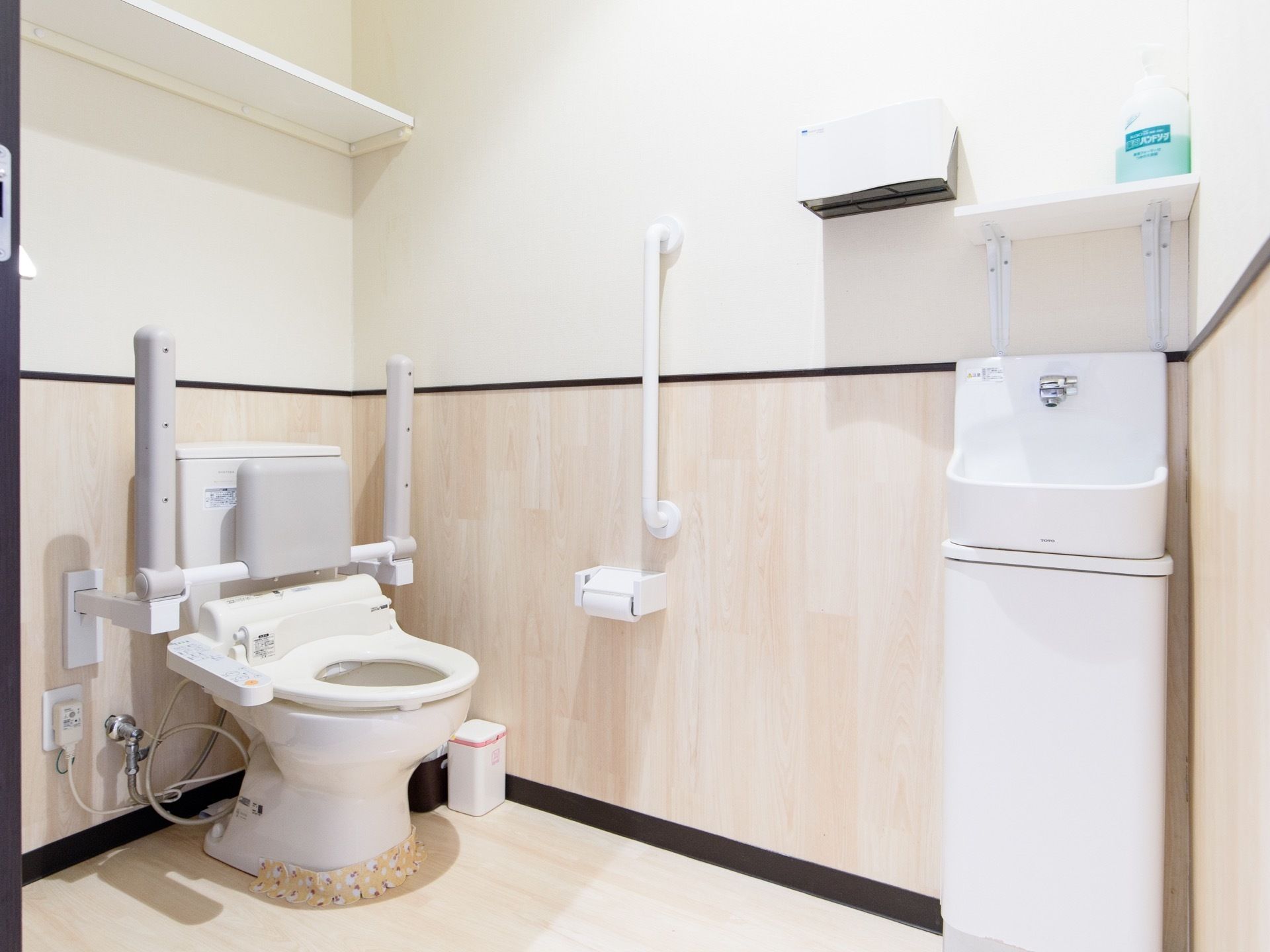 温水洗浄便座を取りつけたトイレで、便座の横に操作パネルがある。便座の両側に折り畳み式の手すりが設置されている。