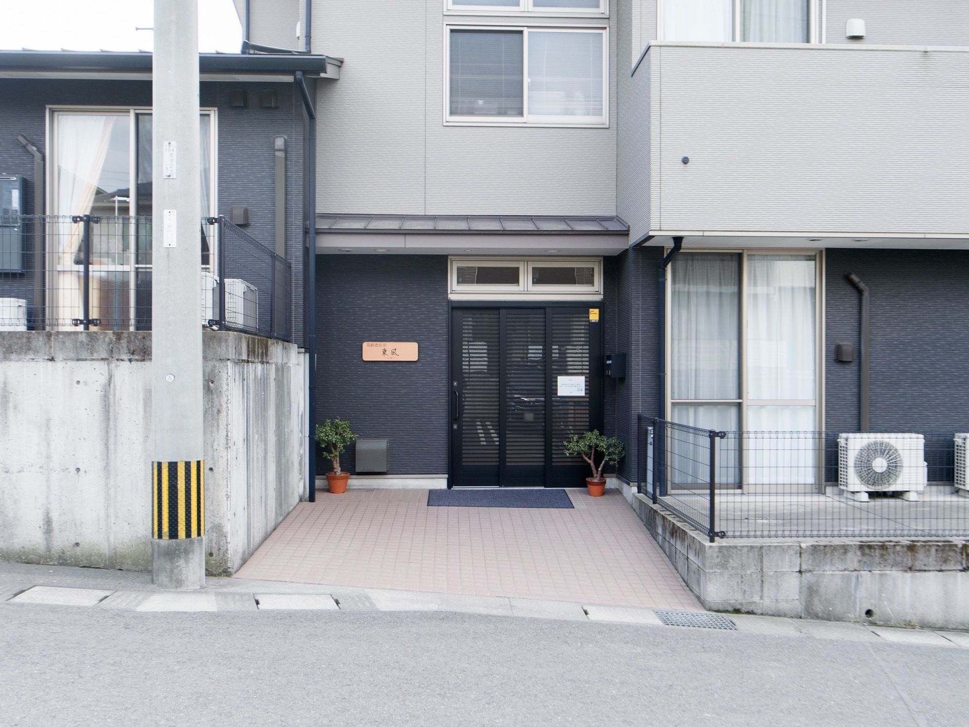 施設前の道路と同じ高さに玄関がある建物である。ダークグレーと薄いグレーに塗り分けられたシックな色合いの建物である。
