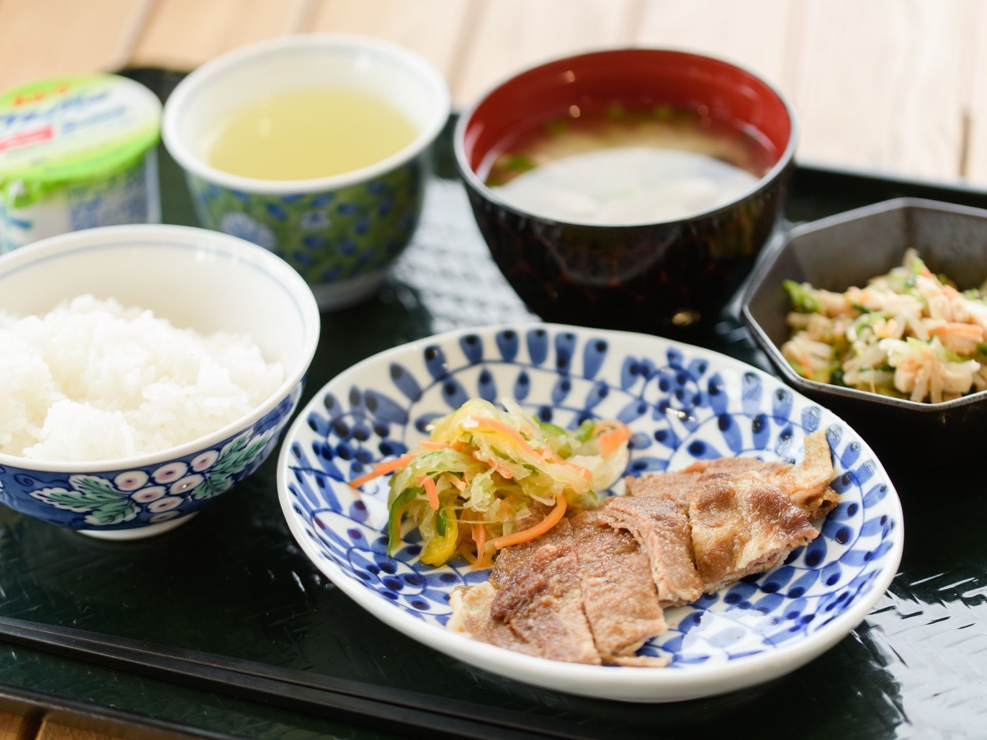黒いトレーに食事がのせられている。白米と味噌汁、焼肉と和え物がメインとなっており、小鉢とデザートもついている。