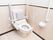 トイレの便座の両側に折り畳み式の手すりが設置されている。温水洗浄便座を採用しており、便座の横に操作パネルがある。