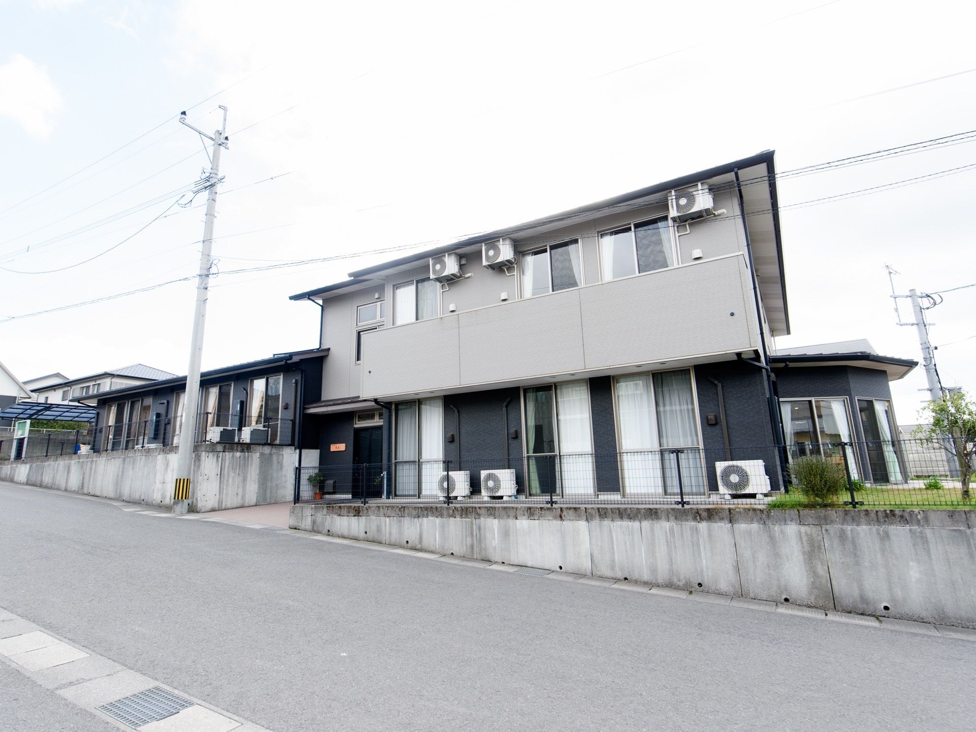薄いグレーの２階建ての建物である。１階部分は黒い外壁で、窓の横に室外機が置かれている。前の通りは緩い上り坂になっている。