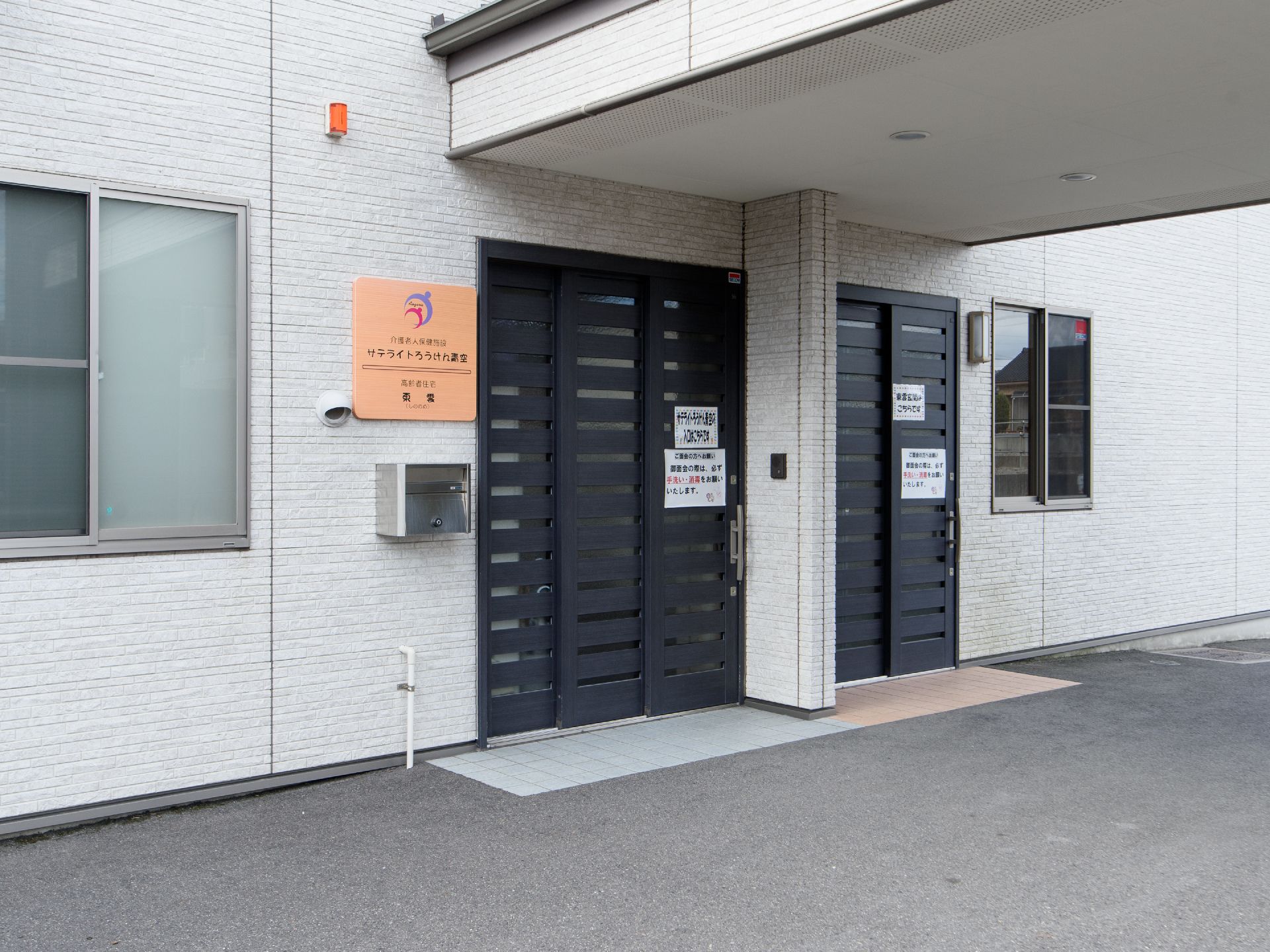壁にホワイトグレーのタイルが貼られた住宅がある。黒い引き戸の玄関で、アプローチはコンクリートで舗装されている。