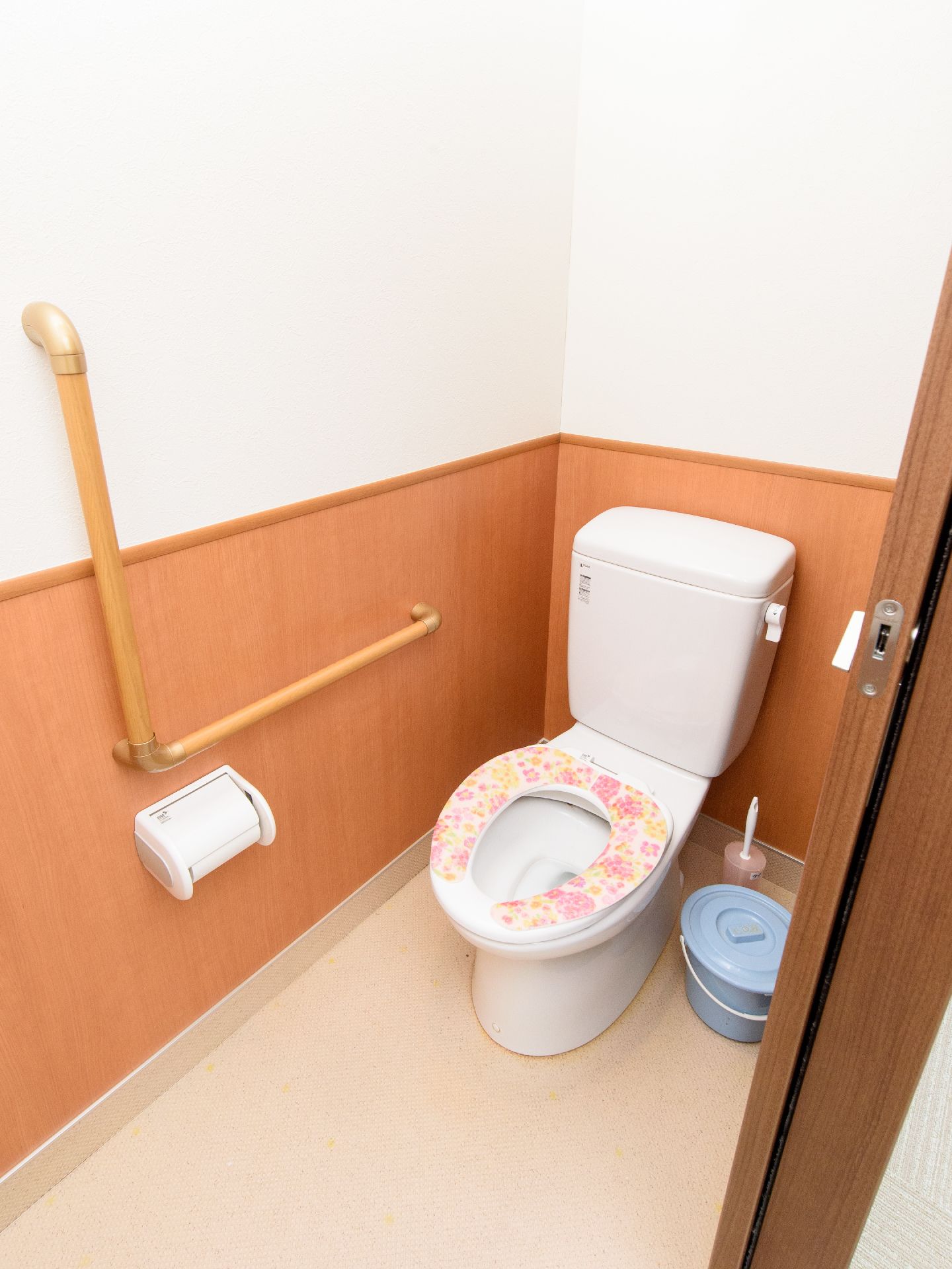 便座に花柄のカバーが掛けられた洋式トイレがある。扉は木製の引き戸で、壁にはL字型の手すりがついている。
