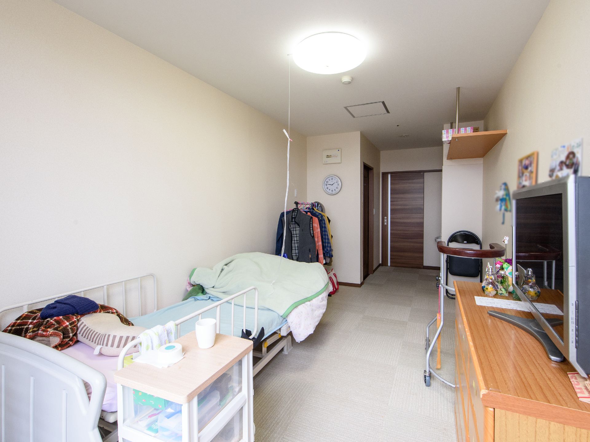カーペット敷きの部屋に、介護用のベッドとチェスト、テレビがある。壁際にはパイプ椅子や歩行器が置かれている。