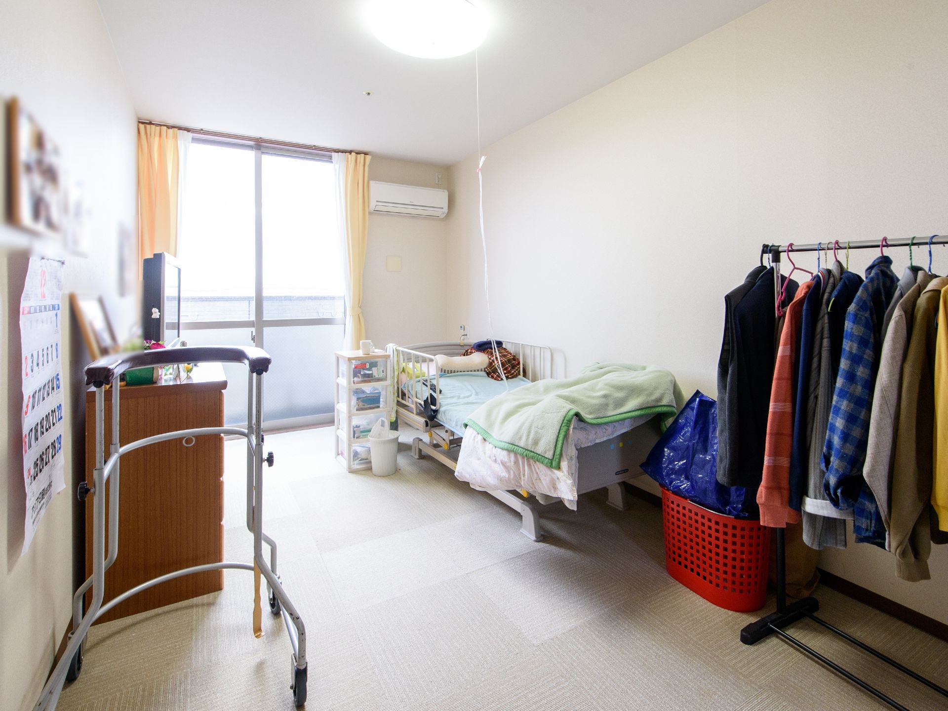 照明とエアコンのついた部屋に、ベッドとハンガーパイプ、タンス、テレビなどが置かれている。カーテンが開いていて、陽が射している。