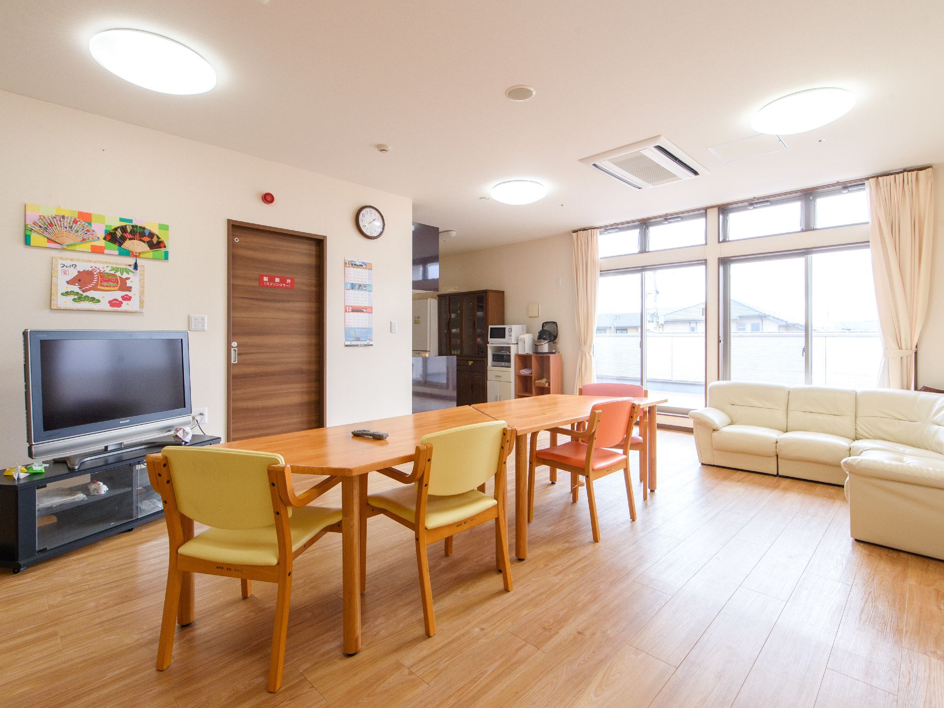 大型テレビが設置されたリビングに、テーブルセットとソファが置かれている。奥にはキッチンがあり、冷蔵庫や食器棚がある。