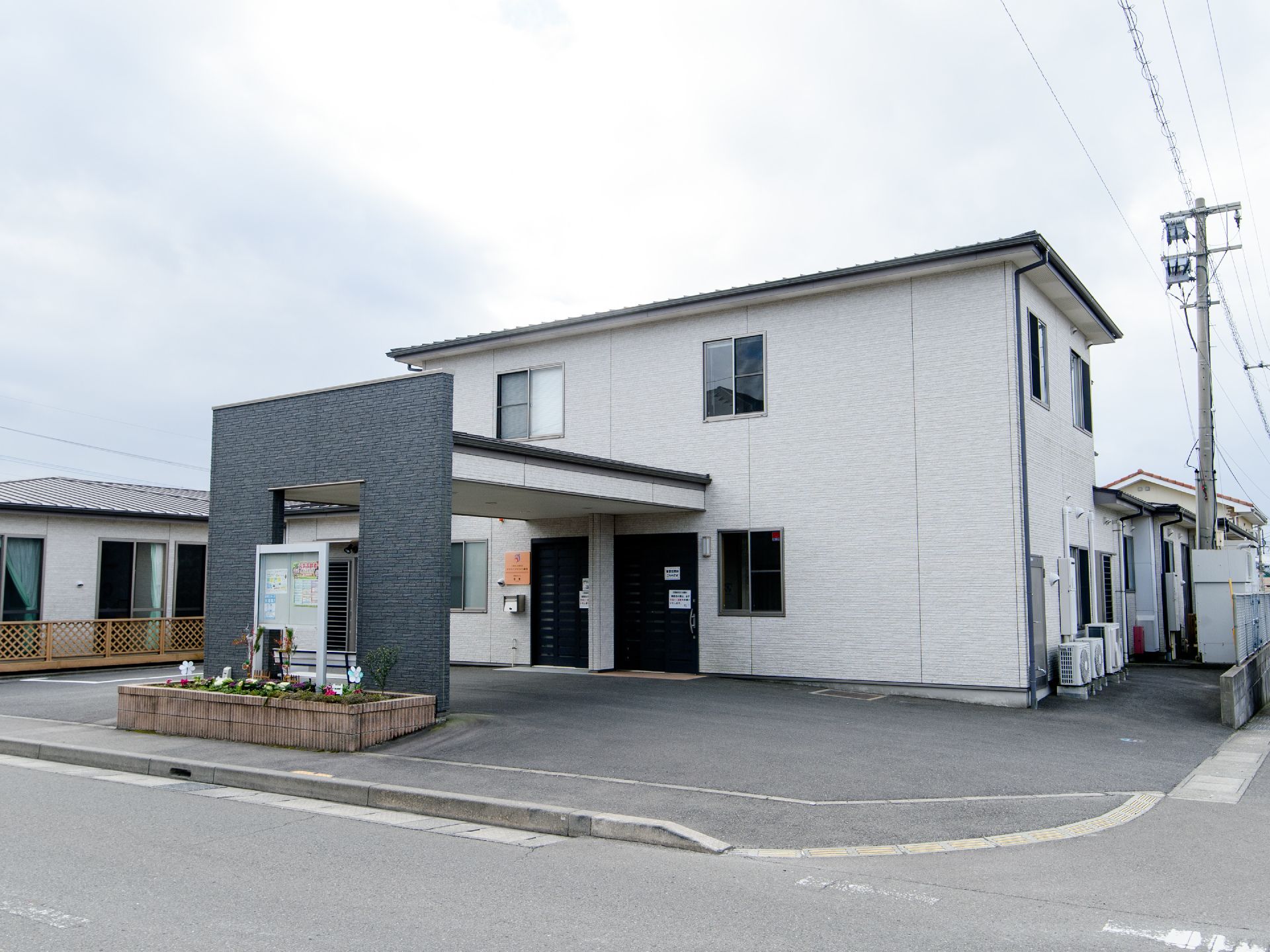 道路沿いの角地に2階建ての住宅が建っている。その周辺は戸建ての多い住宅地になっていて、空が広く見えている。