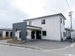 道路沿いの角地に2階建ての住宅が建っている。その周辺は戸建ての多い住宅地になっていて、空が広く見えている。