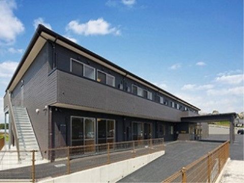 濃いグレーのマンションタイプの建物になっている。玄関の外は屋根になっているため、雨の日の出入りも楽に行える。
