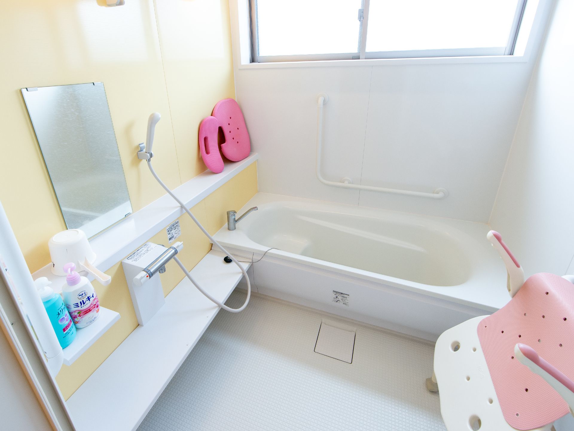 大きな窓のある広い浴室は白とベージュの明るい配色である。シャワーや入浴介助機器があり浴槽にも手すりがついている。