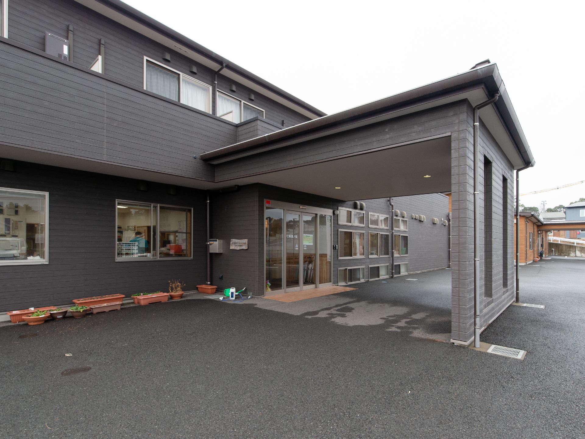 ブラウンの2階建ての横長の建物の端には、ベージュの建物が続いている。玄関には大きなキャノピーが設置され雨天に備えている。