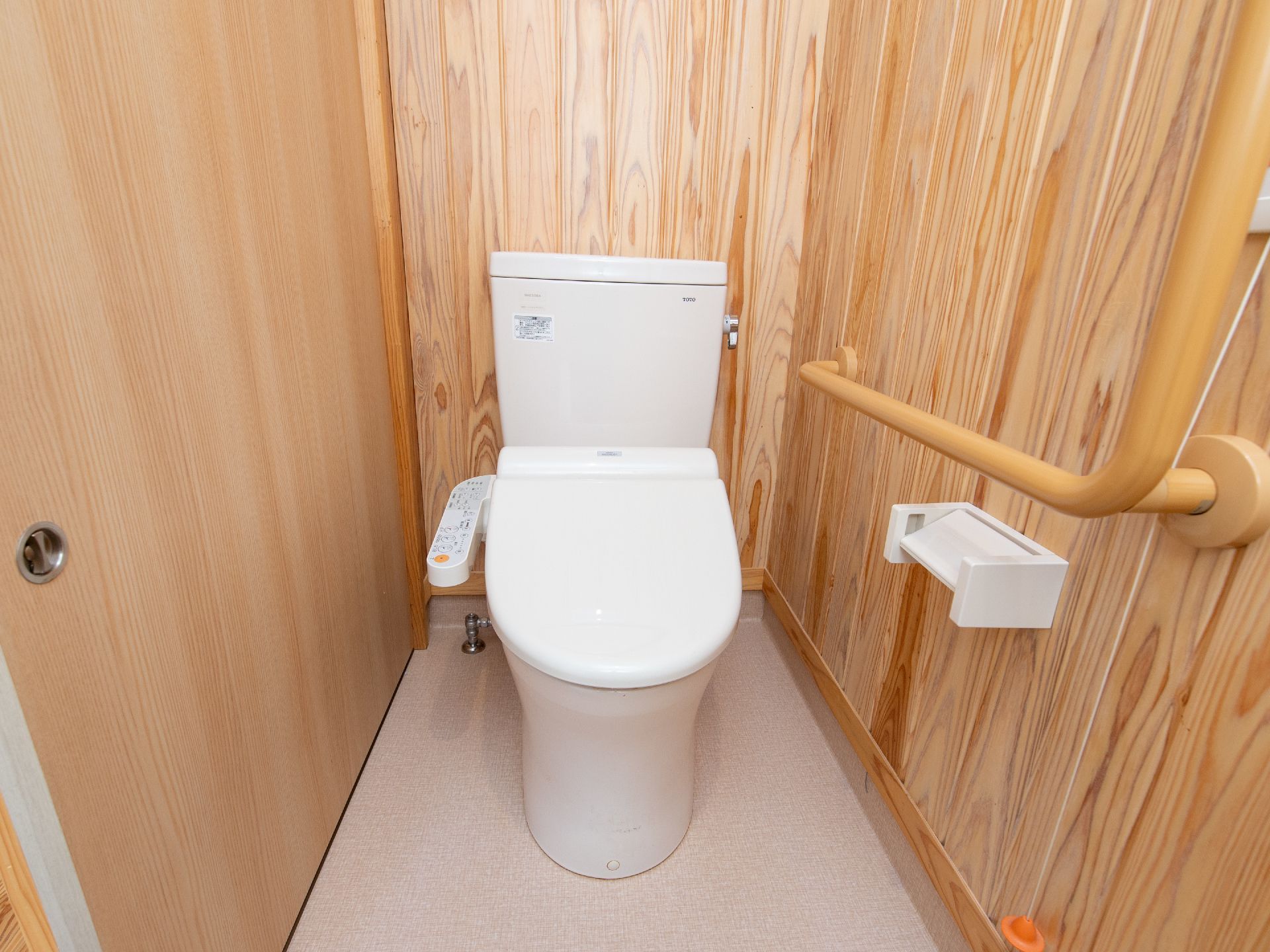 トイレの床は白いフローリングで、温水洗浄の洋式便器が設置されている。木板の壁のペーパーホルダーの上に手すりがついている。