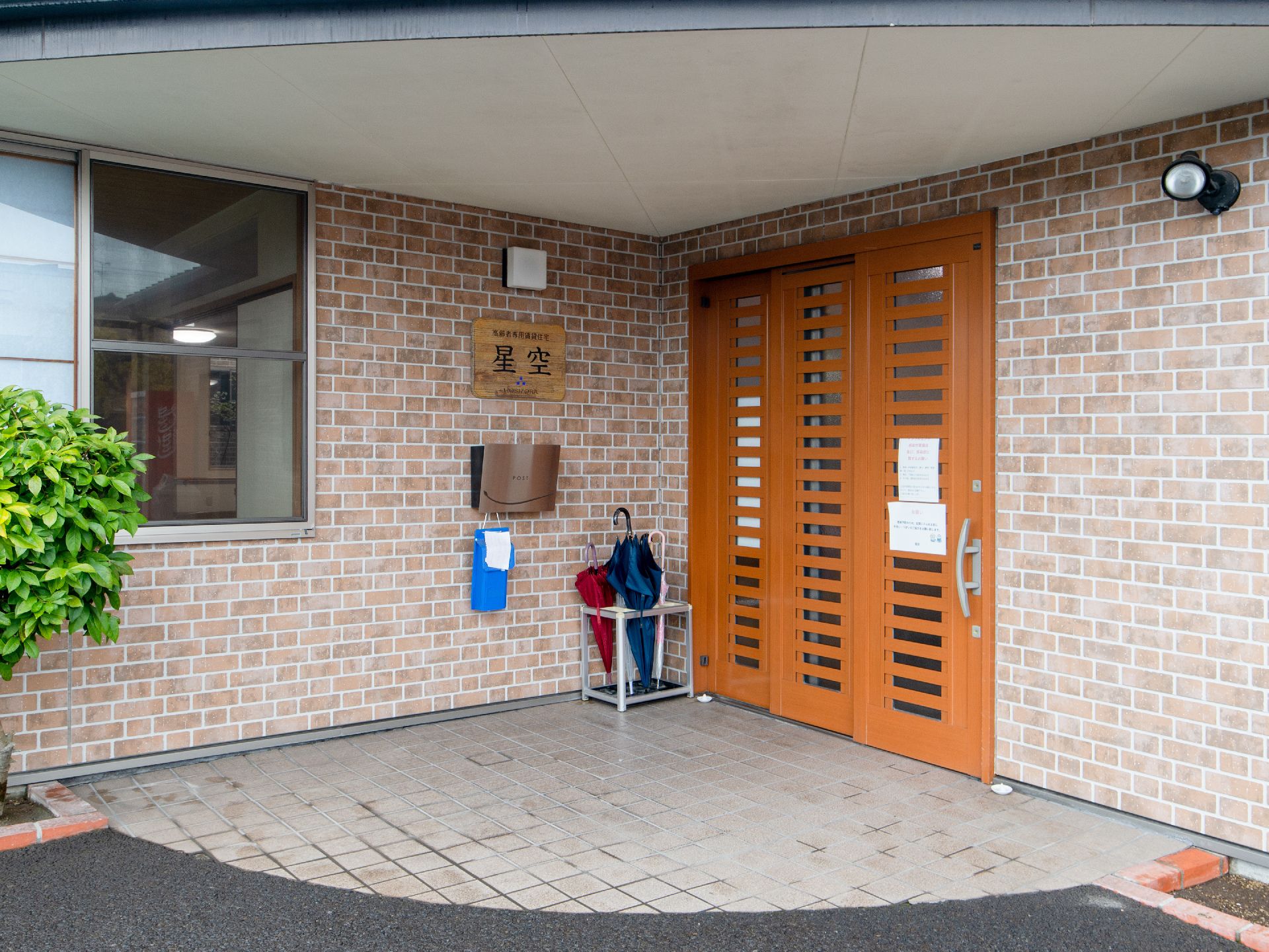大きな屋根を設置した広い玄関である。扉はスライド式になっており、脇には傘立てがある。壁はレンガになっている。