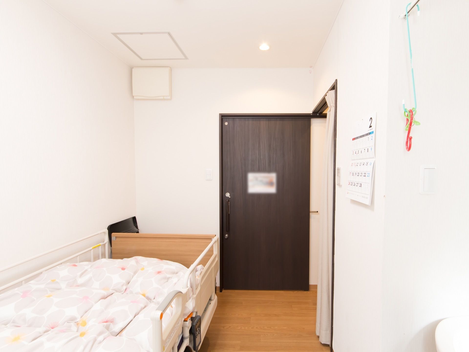ダークブラウンのスライドドアのある居室で、左側にベッドがあり、右側にカーテンで仕切られるトイレが備わっている。