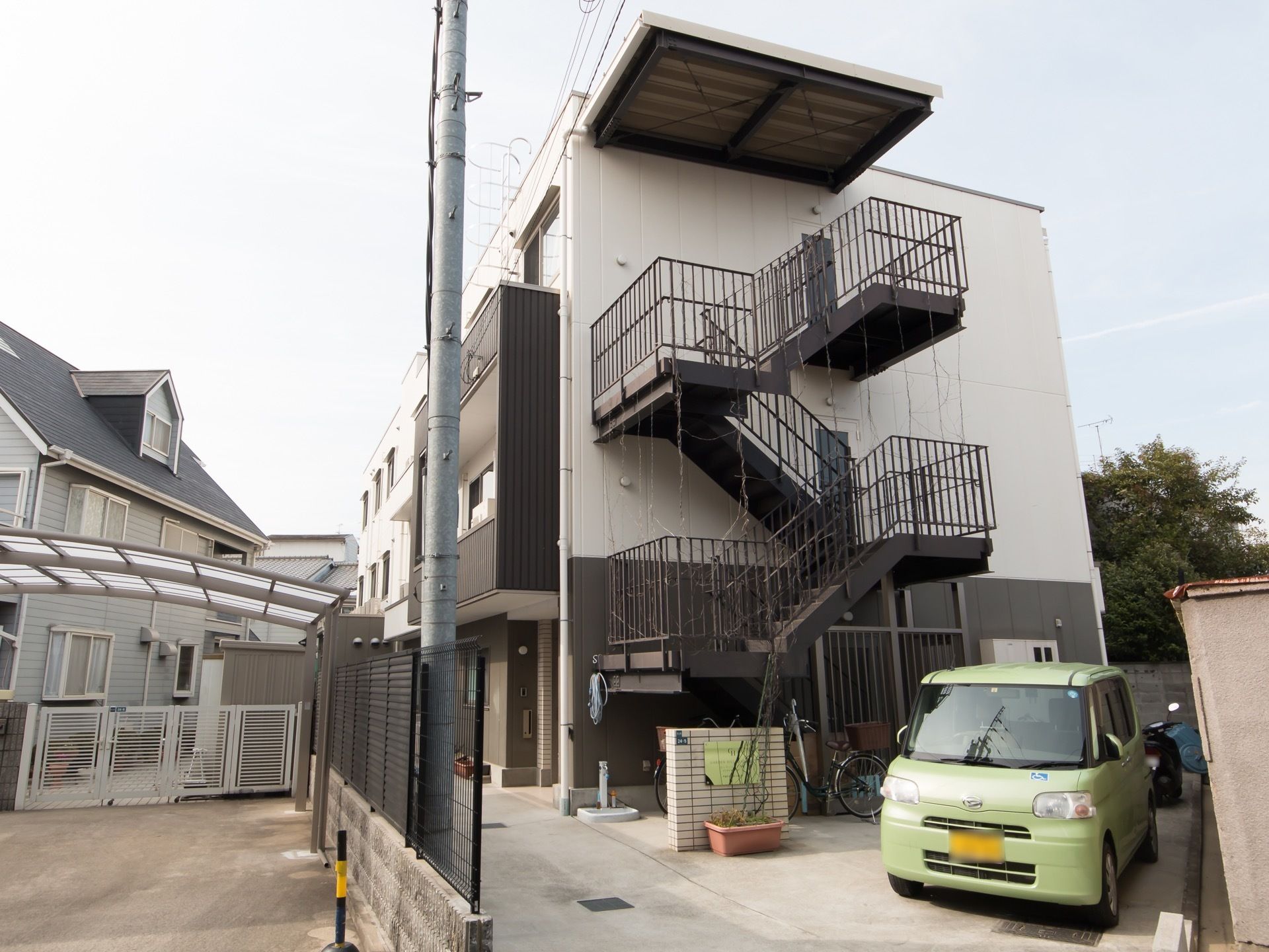 住宅が隣接する施設である。建物前はアスファルトの通路であり、横側のスペースには車や自転車などが停まっている。