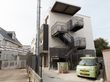 住宅が隣接する施設である。建物前はアスファルトの通路であり、横側のスペースには車や自転車などが停まっている。