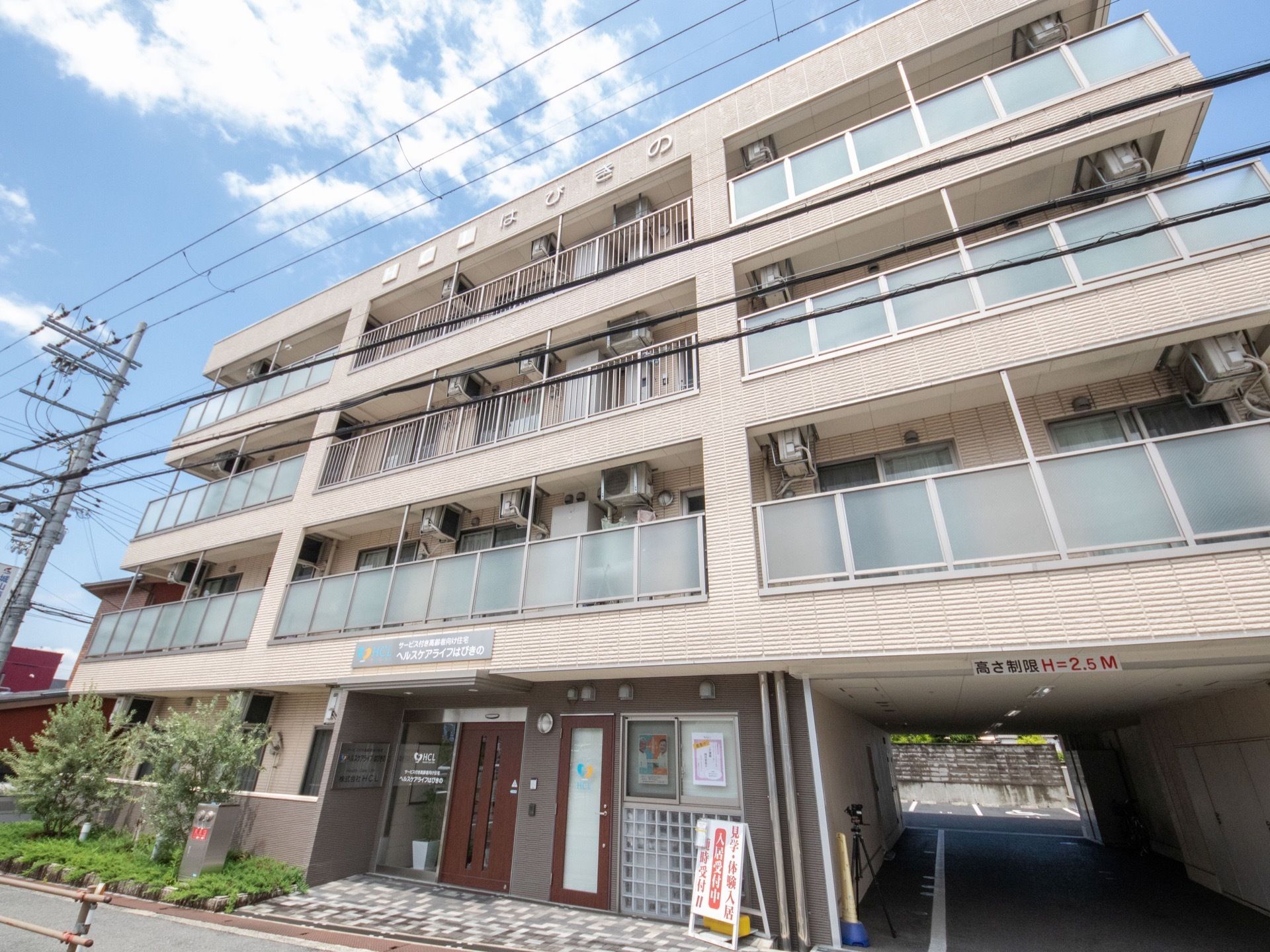 施設は４階建てで、明るい灰色の外壁である。建物の奥には駐車場が見える。施設の入口部には段差の少ない構造になっている。