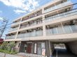 施設は４階建てで、明るい灰色の外壁である。建物の奥には駐車場が見える。施設の入口部には段差の少ない構造になっている。