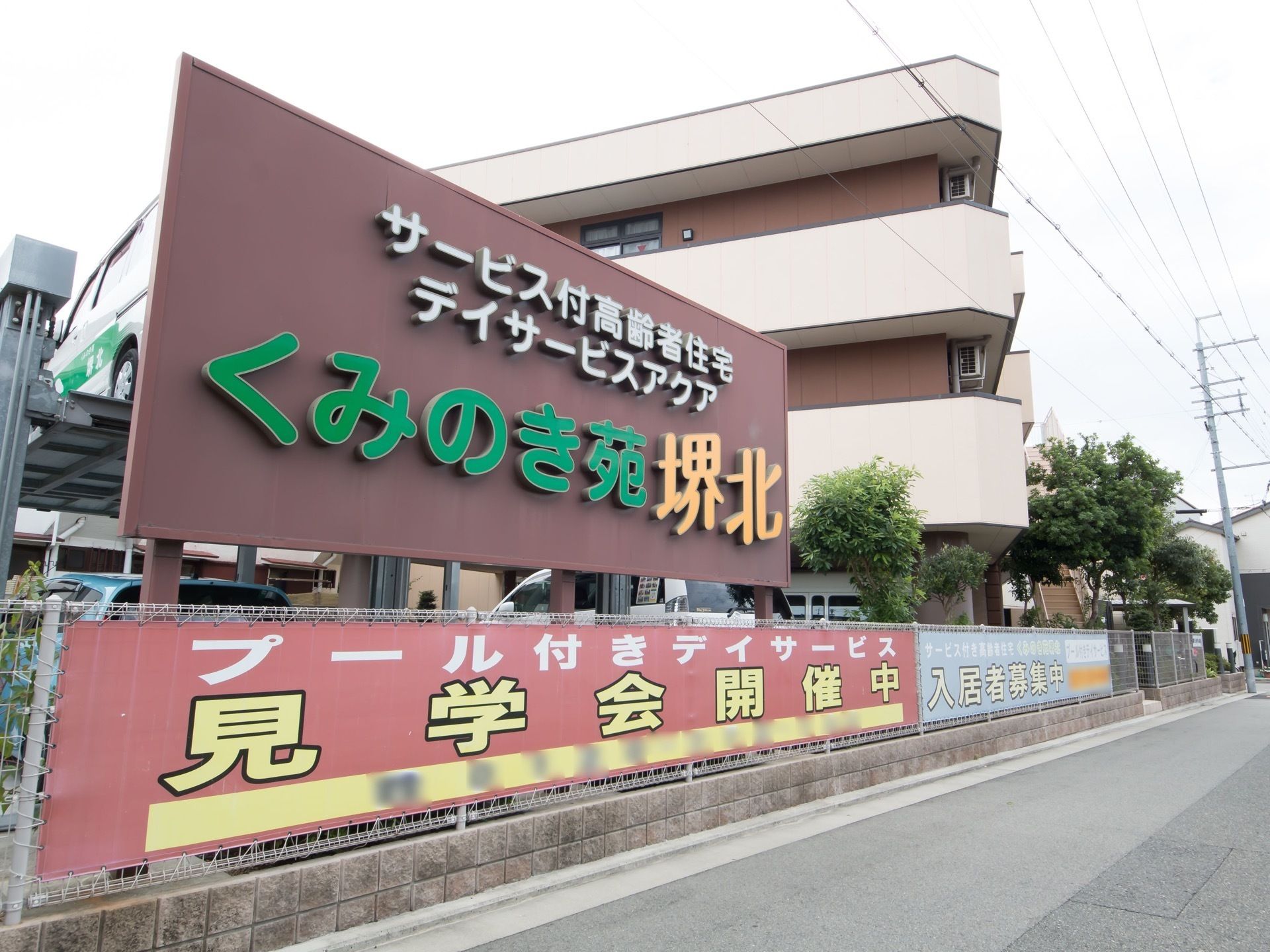 施設名のロゴが入った大きな茶色い看板が、道路に向けて立てられている。支柱は敷地内であるフェンスの内側にある。