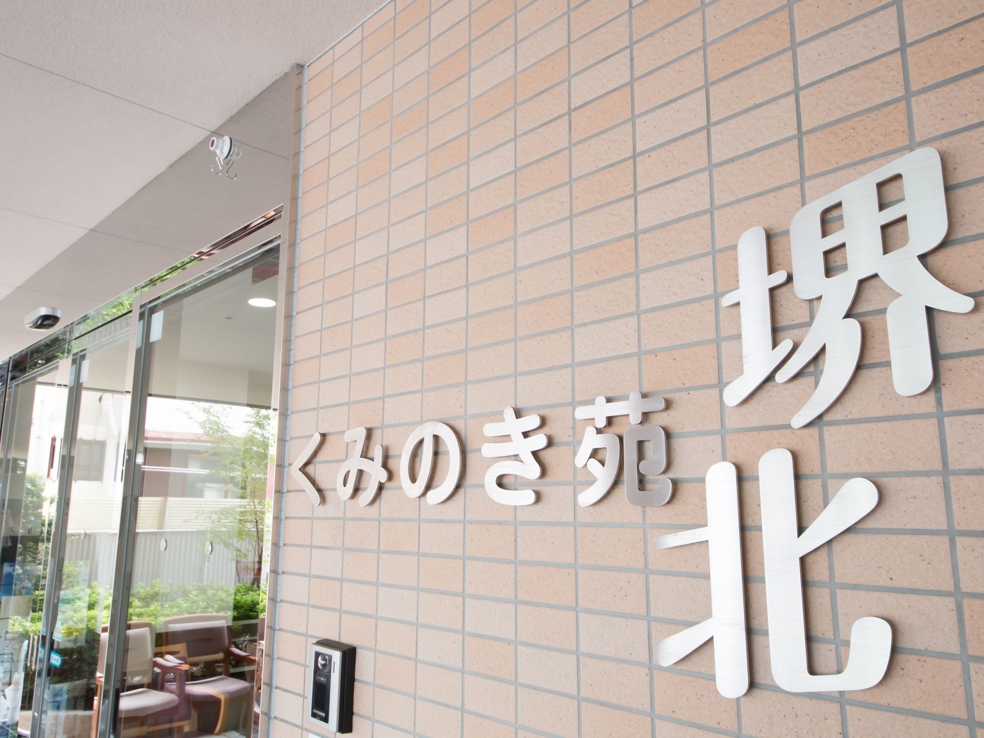 施設の正面玄関の横の壁には、銀色の金属パーツで作られた施設名のロゴが取りつけられている。その下には呼び鈴がある。