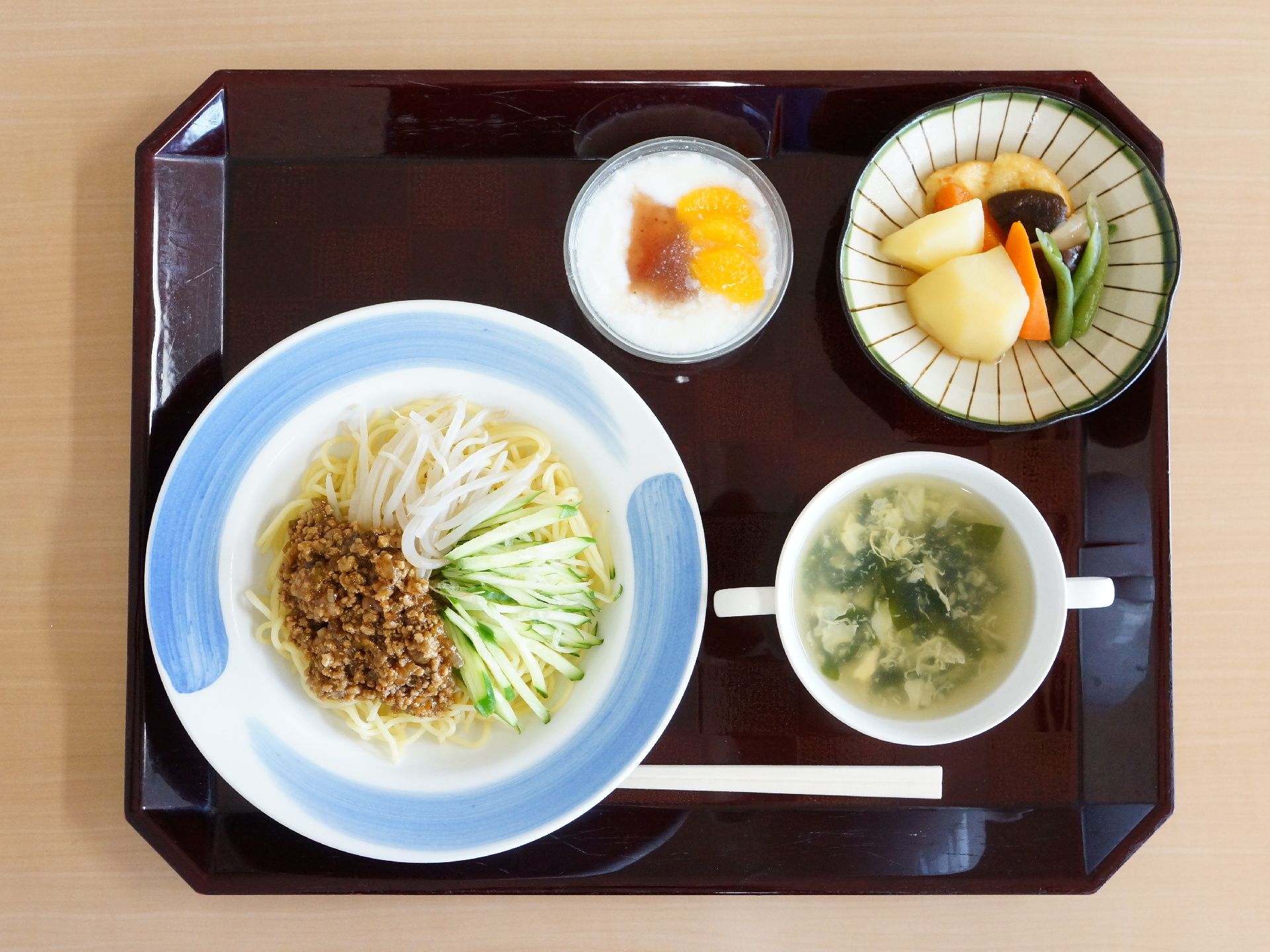 茶色いトレーの上に4つの器と食事が並べられている。きゅうりともやしとひき肉をのせたジャージャー麺とスープ、煮物が盛られている。