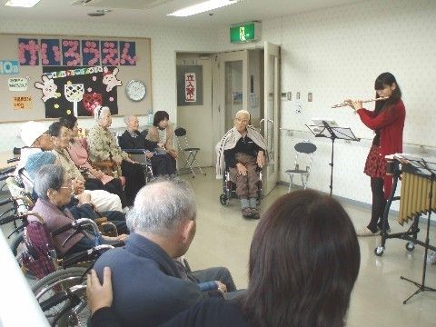 白い壁を背に直立している１人の女性が、楽譜を見ながらフルートを吹いている。周りには車椅子をご利用の入居者様がずらりと並び静かに聴いている。