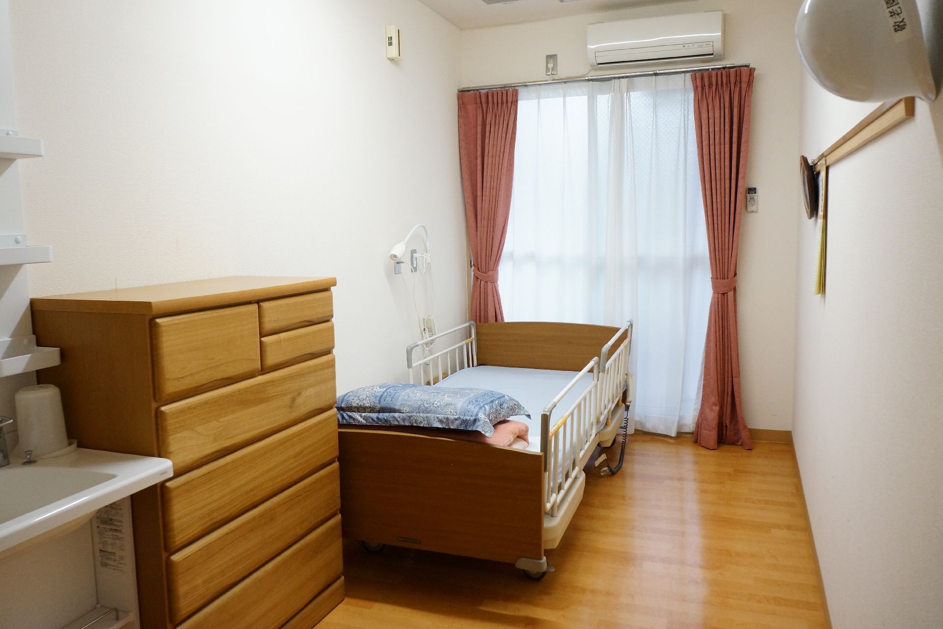 電動ベッド、整理タンス、洗面台が備え付けられた、明るいひとり用の居室