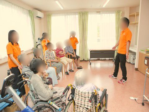 椅子や車椅子に腰かけて、オレンジ色の服を着たスタッフの話を聞いている様子