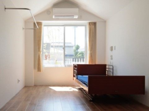 白い壁の居室は、床が茶系のフローリングで、窓の上部にはエアコンが設置されている。壁際にはベッドがあり、壁面にナースコールが付いている。