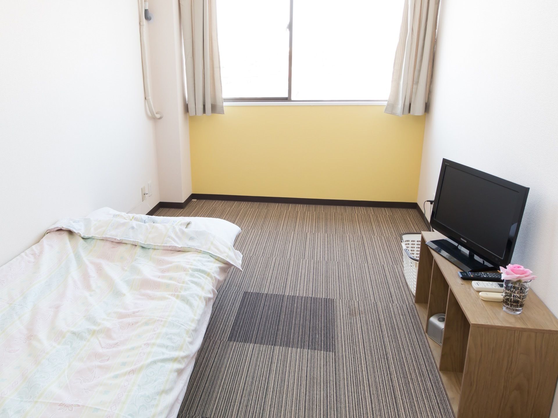 ボーダー柄の絨毯が敷かれた居室で、壁は白と薄い黄色に塗られている。左手の壁際にベッドが置かれている。
