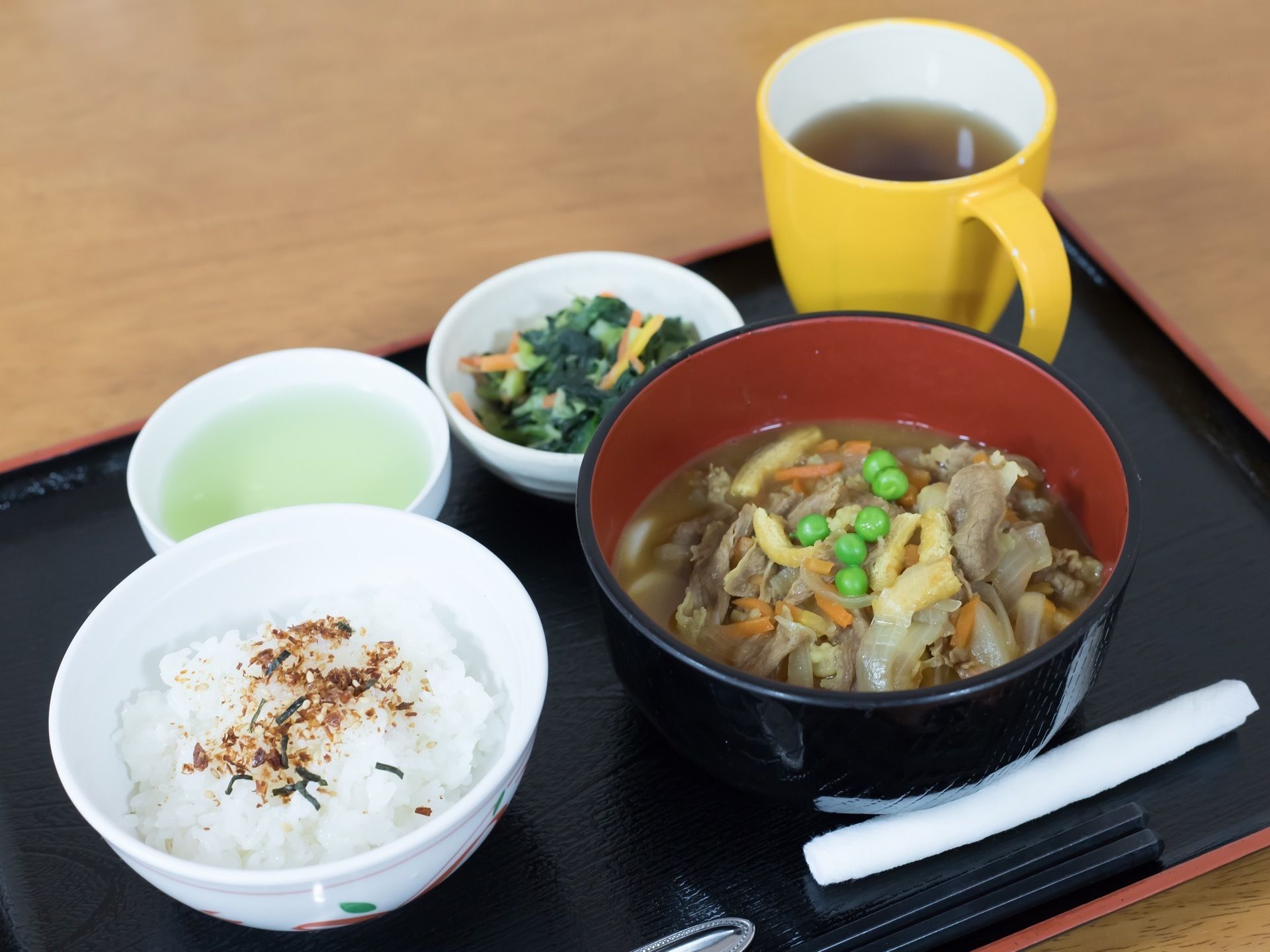 食事は黒い和風のトレイに１人分ずつ配膳されている。白いご飯にふりかけがかかっており、具だくさんの汁物が置かれている。