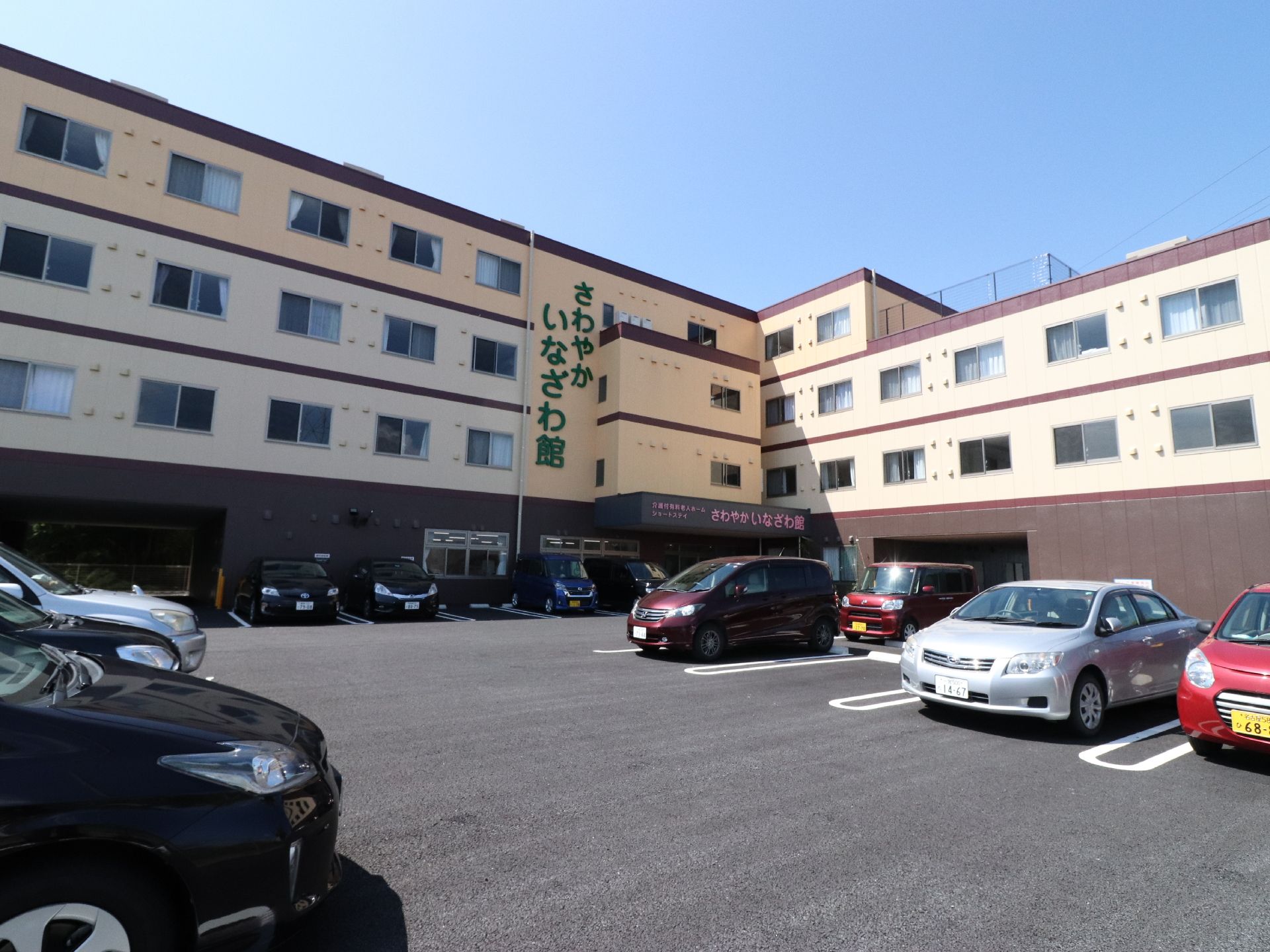 施設の外観。正面に駐車場があり、ベージュとブラウンで彩られた施設が立っている。施設名が大きく書かれた４階建ての建物である。