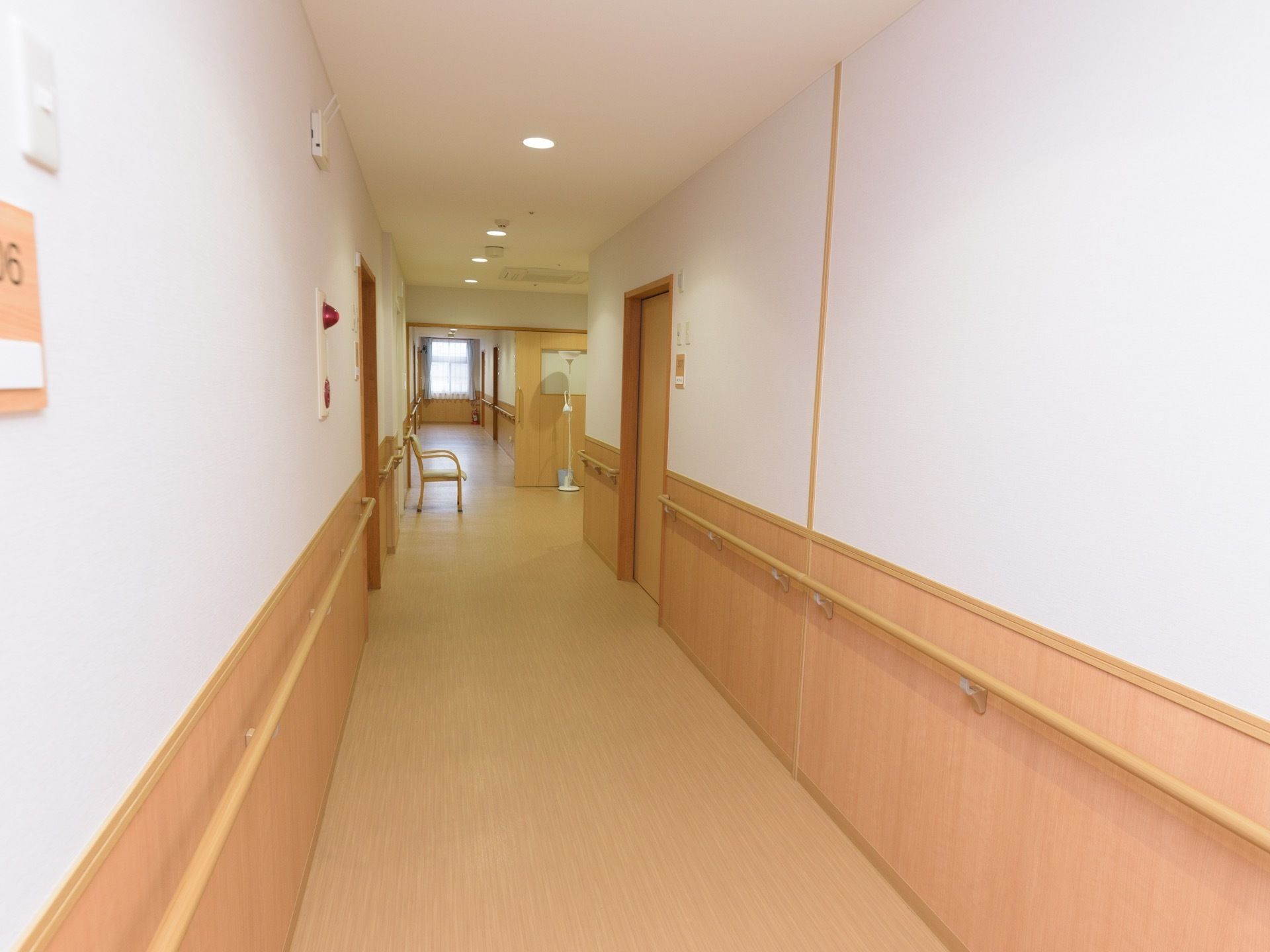 白い壁に明るい木目調を基調に整えられた居室前の廊下である。壁には、手すりが設置され椅子が1脚置かれている。