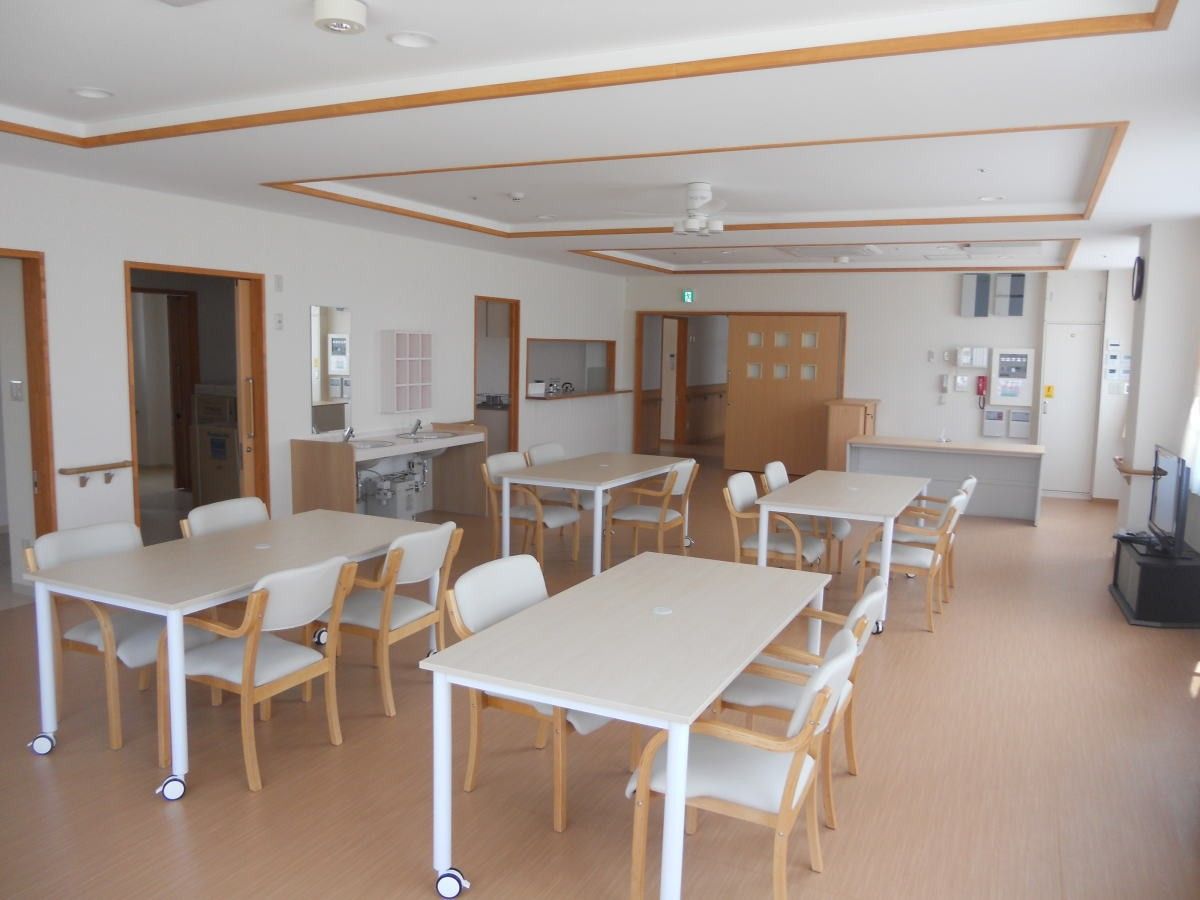 食堂では、入居者様が集いテーブルの席に着かれている。女性スタッフの後ろ姿と数名の車椅子をご利用の方の姿が見える。