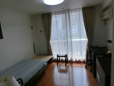 白い壁とフローリングの居室。ベランダに出られる大きな掃き出し窓があり、明るい。ベッドとエアコンが設置されている。