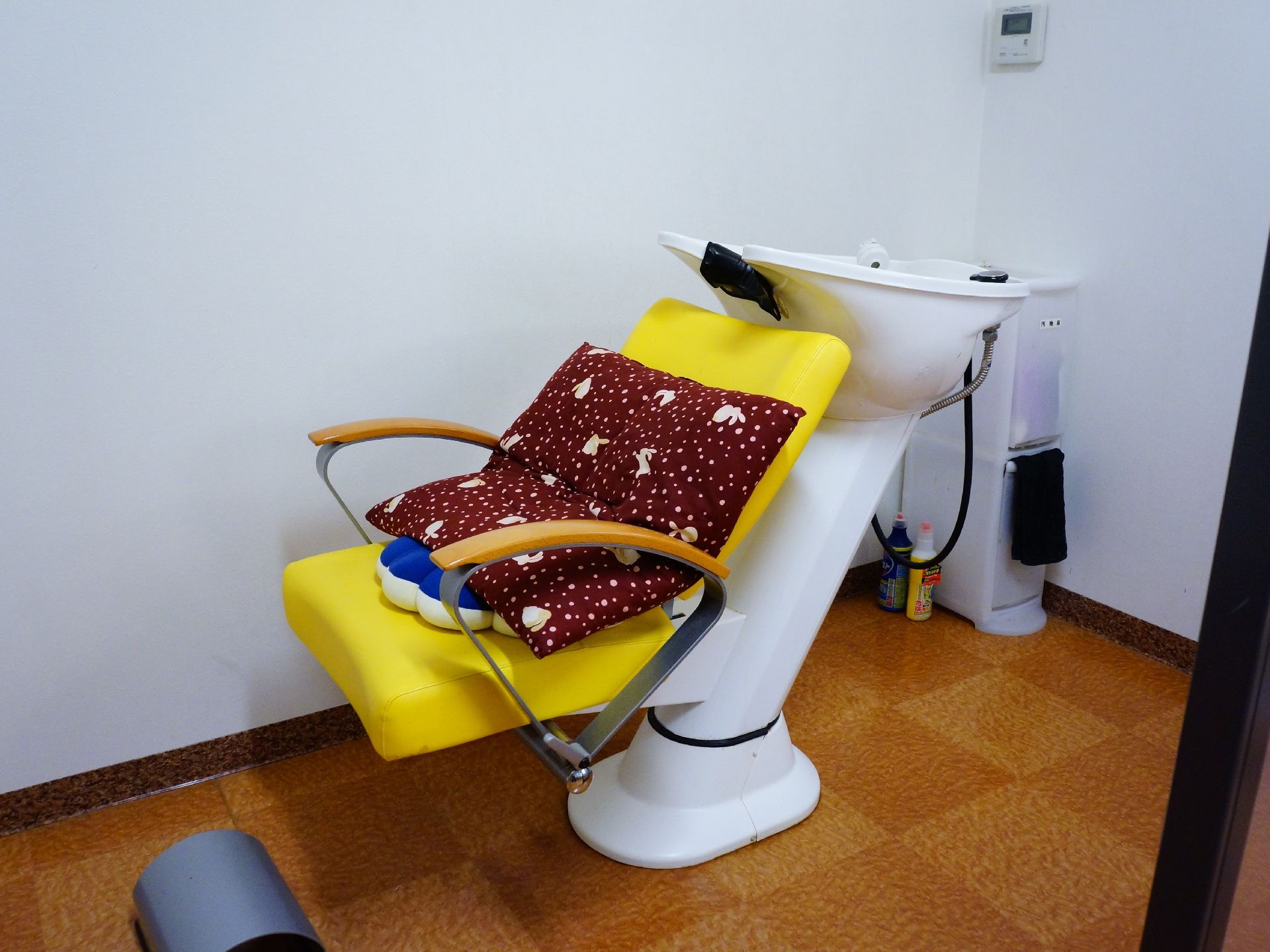 白い壁に茶色のタイルの床の部屋に、黄色のシートの洗髪台があり後ろに頭を倒すと横にならなくても洗えるようになっている。