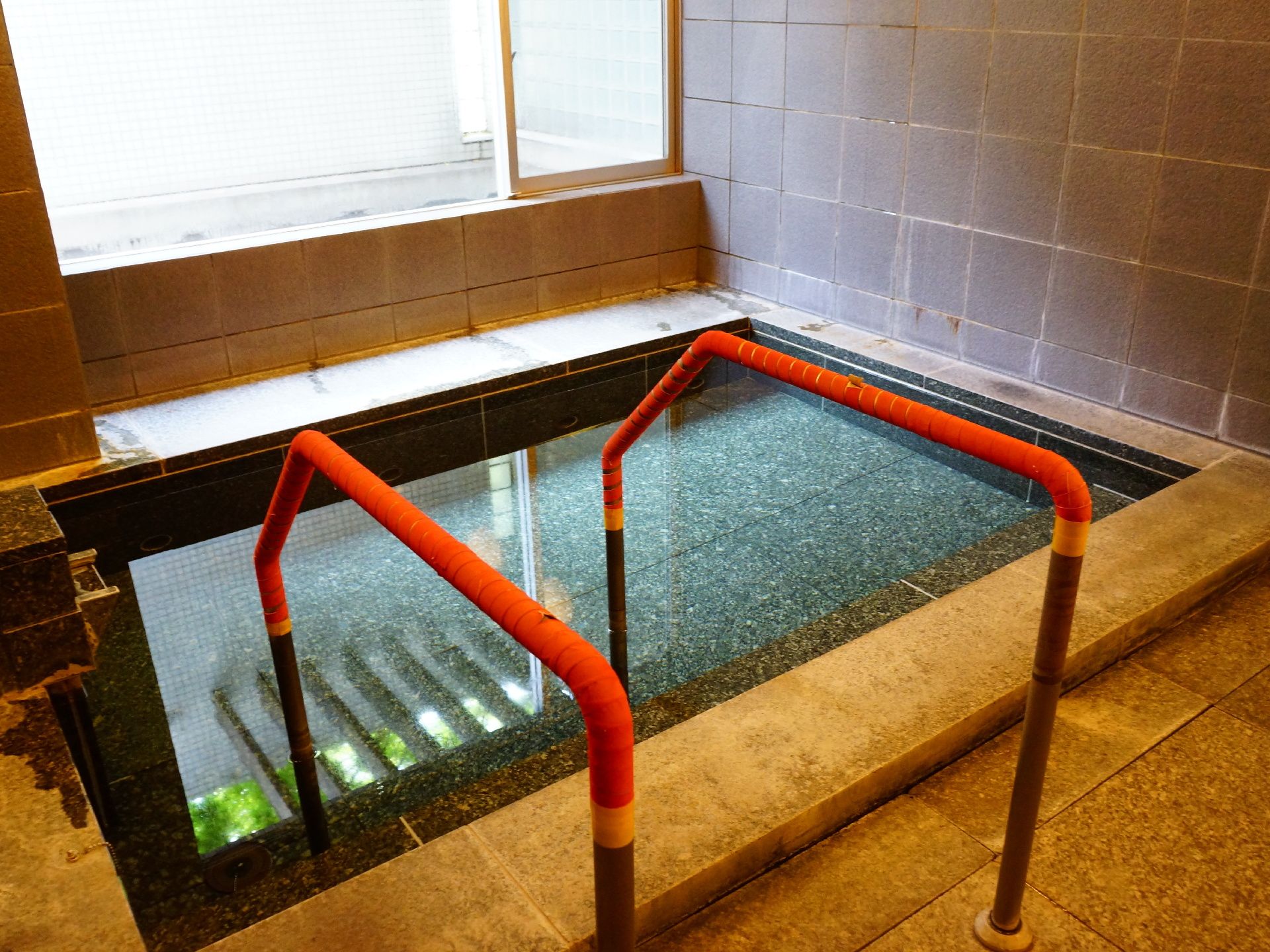 浴槽は窓のすぐ下に位置しており、２本の手すりは水中まで伸びている。壁と床はグレーのタイルが敷き詰められている。